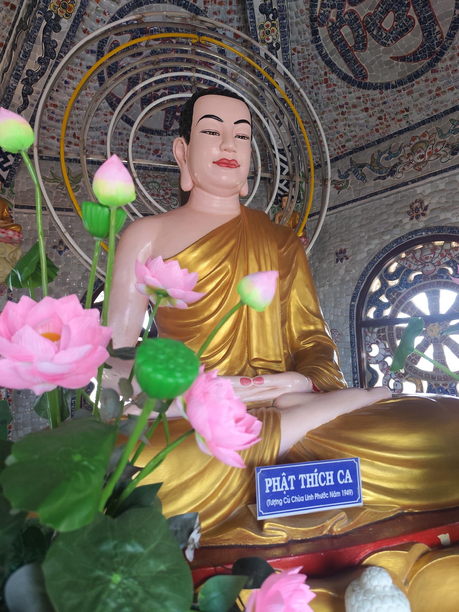 The image shows a golden Buddha statue in a seated meditative pose with pink lotus flowers in the foreground. A sign in Vietnamese, "Phật Thích Ca (Tượng Chùa Linh Phước Năm 1949)", is visible near the base of the statue. The background is decorated with mosaic tiles, and there are circular halo-like structures behind the statue. The image likely originates from a temple, possibly Chùa Linh Phước, located in Xuân Thành, Vietnam.