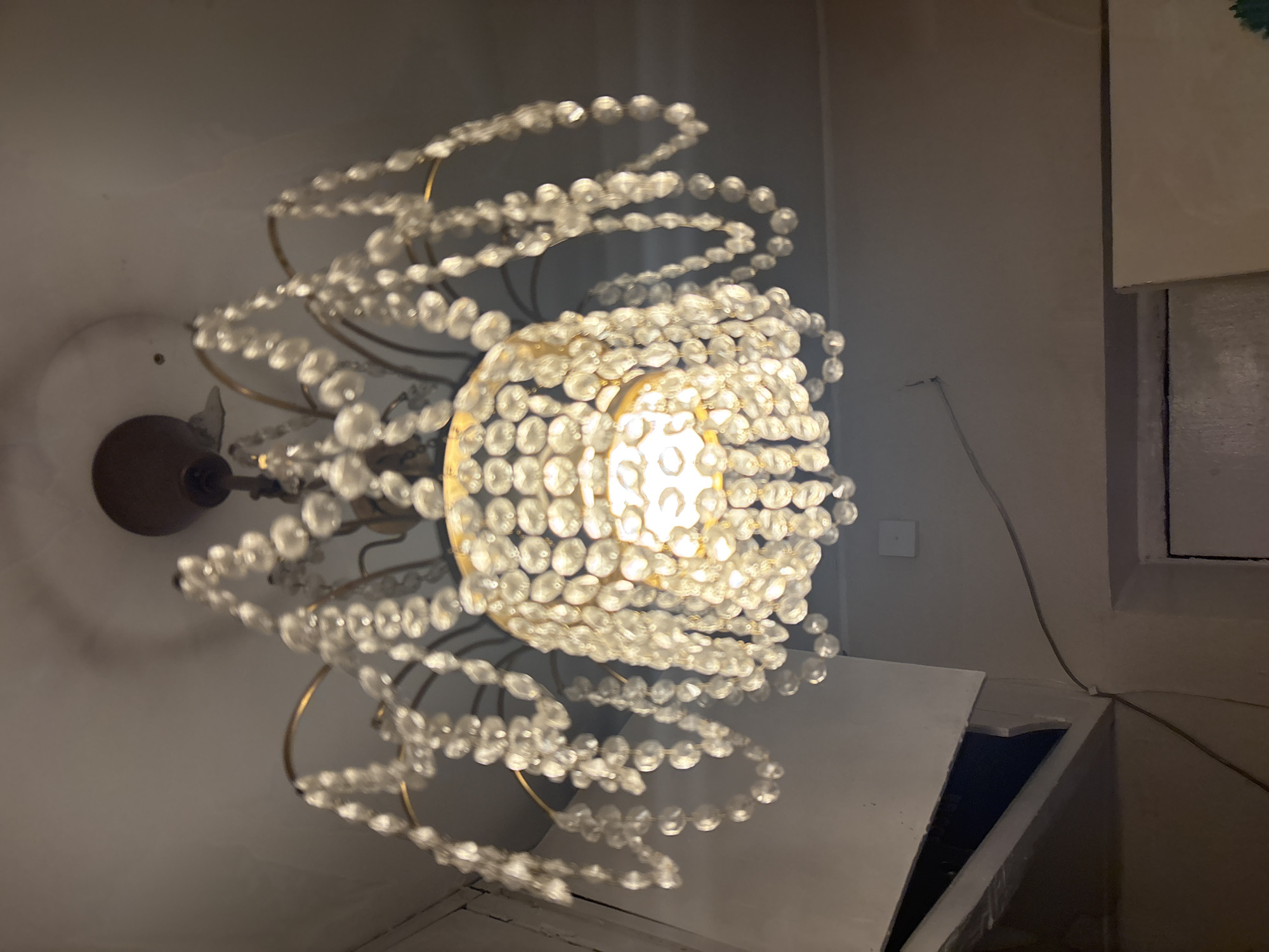 The image shows a close-up, downward-angled view of a crystal chandelier hanging from a ceiling. The chandelier has a metallic frame, likely brass or gold-toned, with multiple tiers of hanging crystal beads. The light bulb within the chandelier is illuminated, casting a warm glow and reflecting off the crystals. The ceiling appears to be white and shows some subtle texture. To the left and bottom of the image, parts of the surrounding room are visible, including a white structure with a dark opening, a portion of a wall, and a thin wire running along the wall. The scene is static and appears to be an indoor shot. No people are present in the image. The location is identified as Lednevo, Russia.