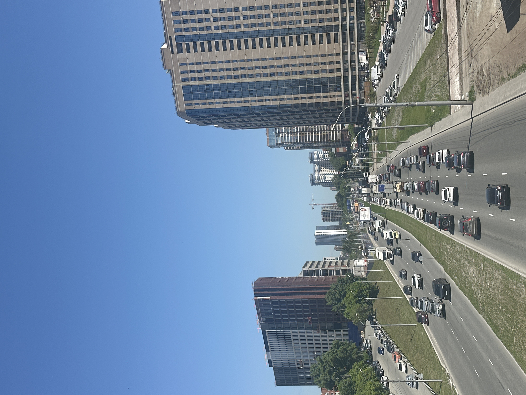This image provides an elevated, wide-angle view of a busy urban highway in Novosibirsk, Russia, on a bright, clear day. The highway, featuring multiple lanes, cuts diagonally across the frame from the foreground towards the horizon, packed with numerous vehicles. Cars, SUVs, and some trucks are visible in both directions, indicating significant traffic flow.
Lining both sides of the expansive road are rows of tall, modern high-rise buildings, interspersed with green grassy areas and trees. On the right side of the highway, a large, multi-story building with numerous windows stands prominently. Further down the road, additional high-rises are seen, some partially obscured by lush green foliage. On the left side, a section of the city showcases a mix of completed buildings and one particularly large structure under active construction, with visible frameworks and scaffolding. A construction crane can be spotted in the distance, underscoring ongoing urban development.
The sky is a clear, vibrant blue, characteristic of a sunny day, likely midday or early afternoon given the bright illumination and distinct shadows. The scene captures the dynamic atmosphere of a large city, focusing on its transportation infrastructure and dense architectural landscape. No individuals are distinctly visible due to the elevated perspective, and any text on distant billboards is illegible.