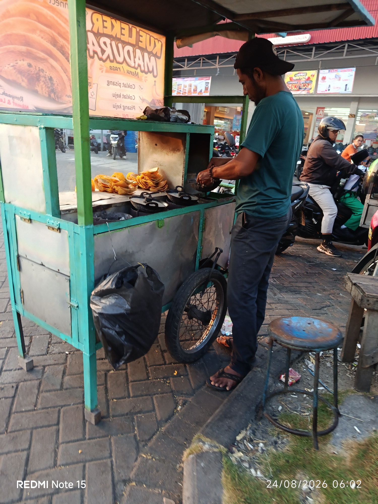 In Keboansikep, Indonesia, an outdoor food stall is depicted. A man in a teal shirt and dark pants stands behind the stall, actively preparing food. The stall itself is constructed with a turquoise metal frame and features a stainless steel countertop. On the counter, several small black pans are visible, along with a pile of golden-brown, circular food items. Behind the pans, a stack of similar food items is arranged. A large black plastic bag hangs from the side of the stall. To the left, a wheel with visible spokes is attached to the stall. In the foreground, a weathered blue and black stool sits on the paved ground. The background shows a street scene with motorcycles and people, suggesting a public area or market. The overall lighting indicates it is daytime, possibly late afternoon or early evening given the warm tones.