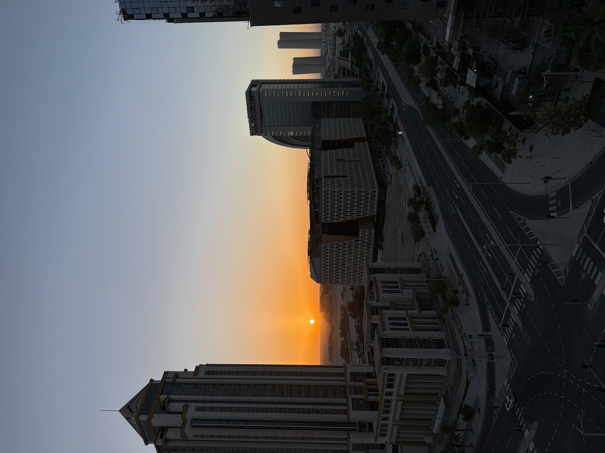 This high-angle shot captures a city scene at sunset. The sun, a bright orange orb, is visible near the horizon, casting a warm glow across the sky and reflecting off the windows of the surrounding skyscrapers. The architecture is modern, with sleek glass buildings dominating the skyline. A multi-lane road with a few cars is visible in the foreground, curving through the urban landscape. The sky transitions from a pale blue at the top to a warm orange and yellow hue near the horizon. The scene appears to be an urban development with ongoing construction visible in some areas. The overall impression is one of a dynamic and growing city bathed in the beautiful light of sunset.