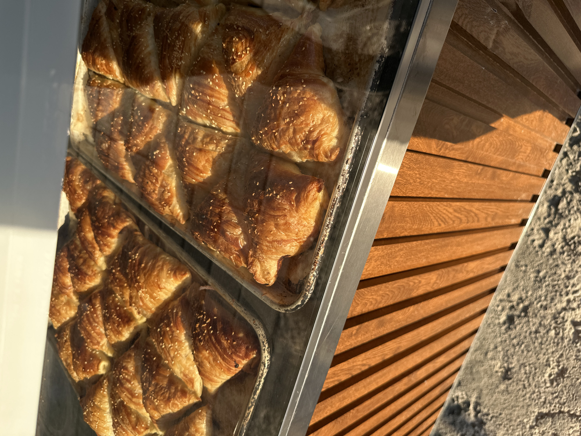 The image shows two baking trays filled with golden-brown pastries, possibly croissants or samsa, dusted with sesame seeds. The pastries are arranged in neat rows and appear to be fresh. The trays are placed on a counter behind a glass display case. The counter itself is made of wooden slats with a textured, sandy surface below. The scene suggests a bakery or a food stall. The lighting indicates it is either early morning or late afternoon, with sunlight casting shadows across the wooden surface. The specific location in Tashkent, Uzbekistan cannot be determined from the image alone.