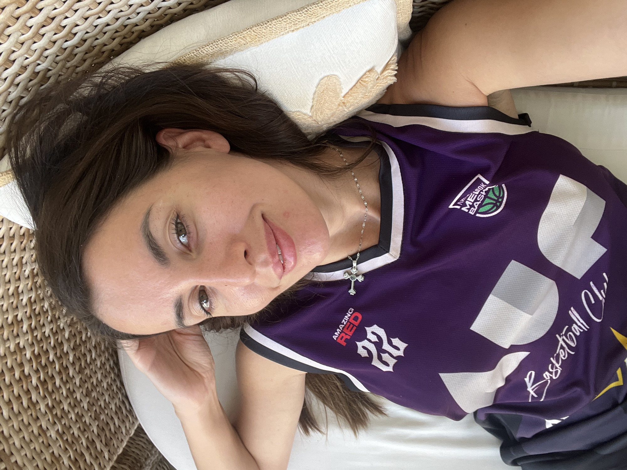 A woman with dark brown hair is lying down on a cushioned surface, possibly a bed or lounger. She is wearing a purple basketball jersey with white and black trim, and a silver necklace with a cross pendant. Her right hand is resting on the back of her head, and she is looking at the camera with a slight smile. The background shows a woven texture, likely part of the furniture. There is no specific identifiable information within the image to pinpoint a location in North Kuta, Indonesia.