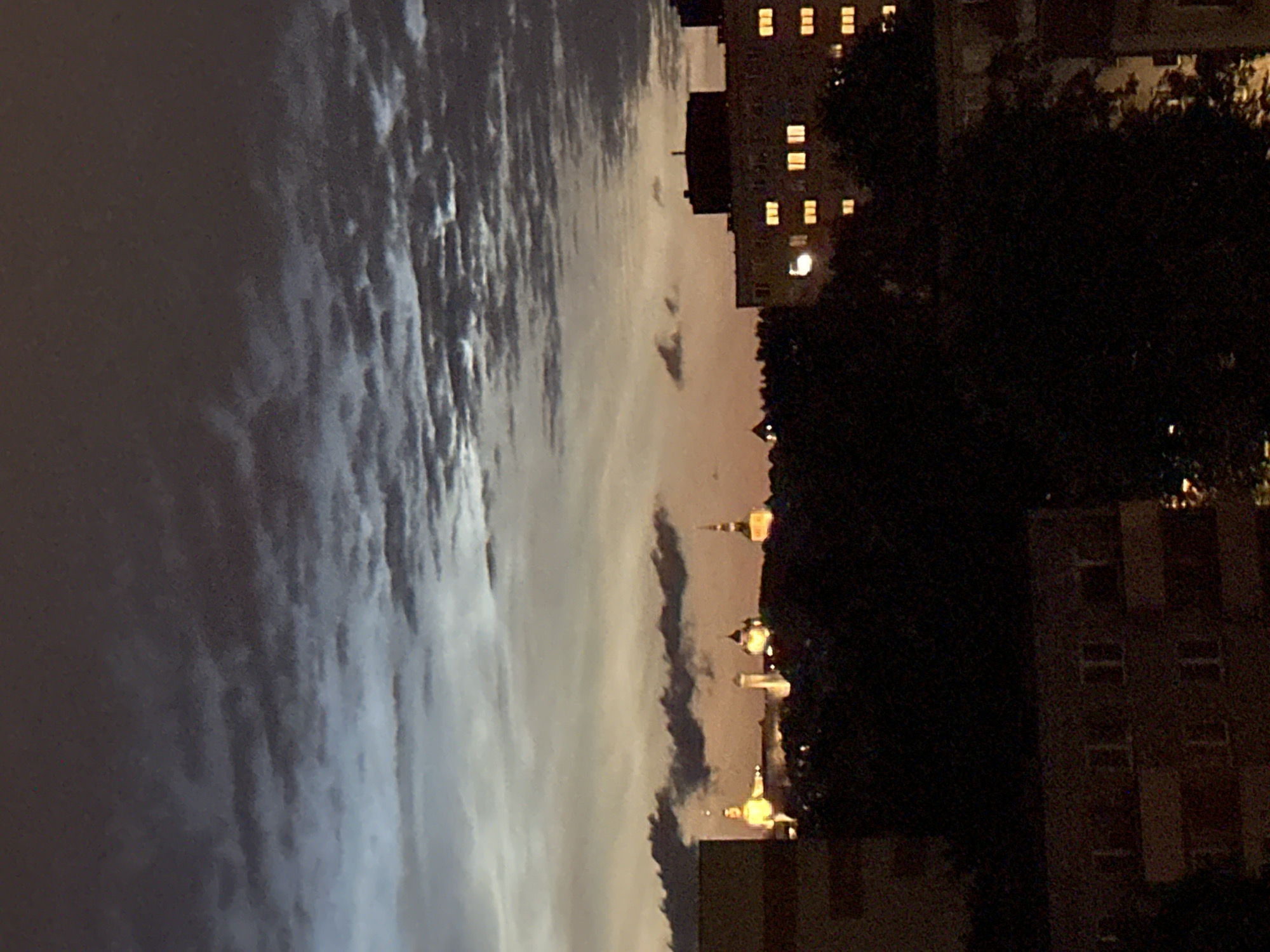 The image depicts a nighttime cityscape, likely in Tallinn, Estonia, characterized by a gradient of sky colors from dark gray clouds at the top to a soft orange and blue hue near the horizon. Several buildings are visible, with illuminated windows suggesting occupancy. In the middle ground, a silhouette of trees partially obscures a skyline featuring several illuminated spires and domes, indicative of historical architecture or religious structures. To the right, a large apartment building with numerous lit windows stands out. The foreground is dominated by dark shapes of buildings and trees, rendered almost entirely in shadow. The overall scene conveys a sense of urban twilight or early night.