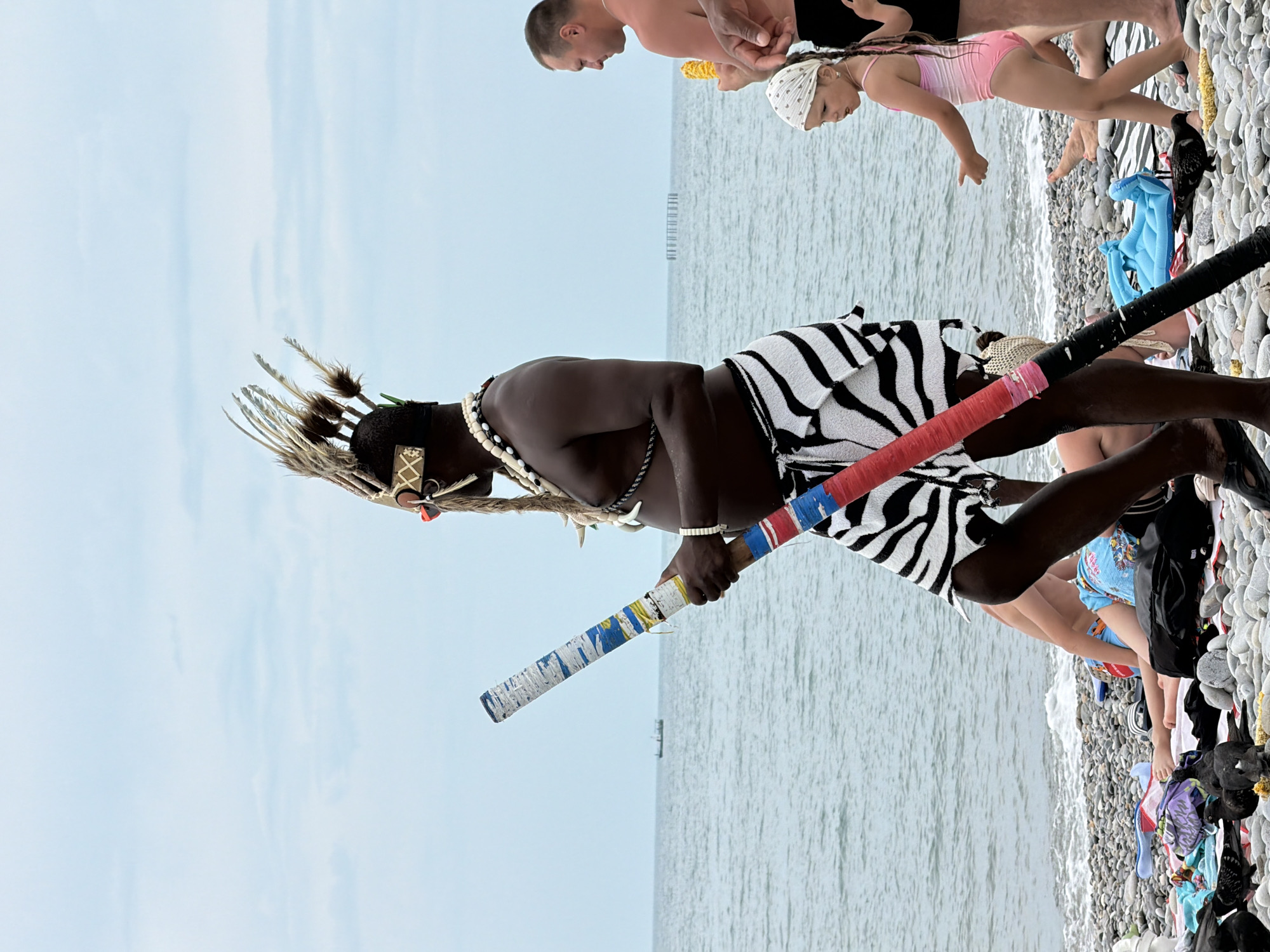 The image depicts a group of people on a pebble beach with the sea in the background. The scene is oriented vertically, with the sky at the top and the sea and beach at the bottom.
The main subject in the foreground is a dark-skinned person wearing a traditional outfit consisting of a black and white striped garment, a beaded necklace, and a headdress adorned with feathers and animal teeth. This person is holding two decorated staffs, one with red, blue, and white bands, and the other with a red band.
In the background, several other individuals are visible. A man and a young girl, possibly his daughter, are standing closer to the sea. The girl is wearing a pink swimsuit and a white bandana. Other people are sitting or standing on the beach, some partially obscured by the main subject or other objects. Towels and bags are scattered on the pebbles.
The activity appears to be a casual gathering at the beach, possibly with a performance or presentation by the person in traditional attire. The weather seems overcast, indicated by the muted light and the appearance of the sky. The presence of the sea and pebbles strongly suggests a coastal location, consistent with the provided context of Sochi, Russia. The overall mood is relaxed and informal. No visible text is present in the image.
