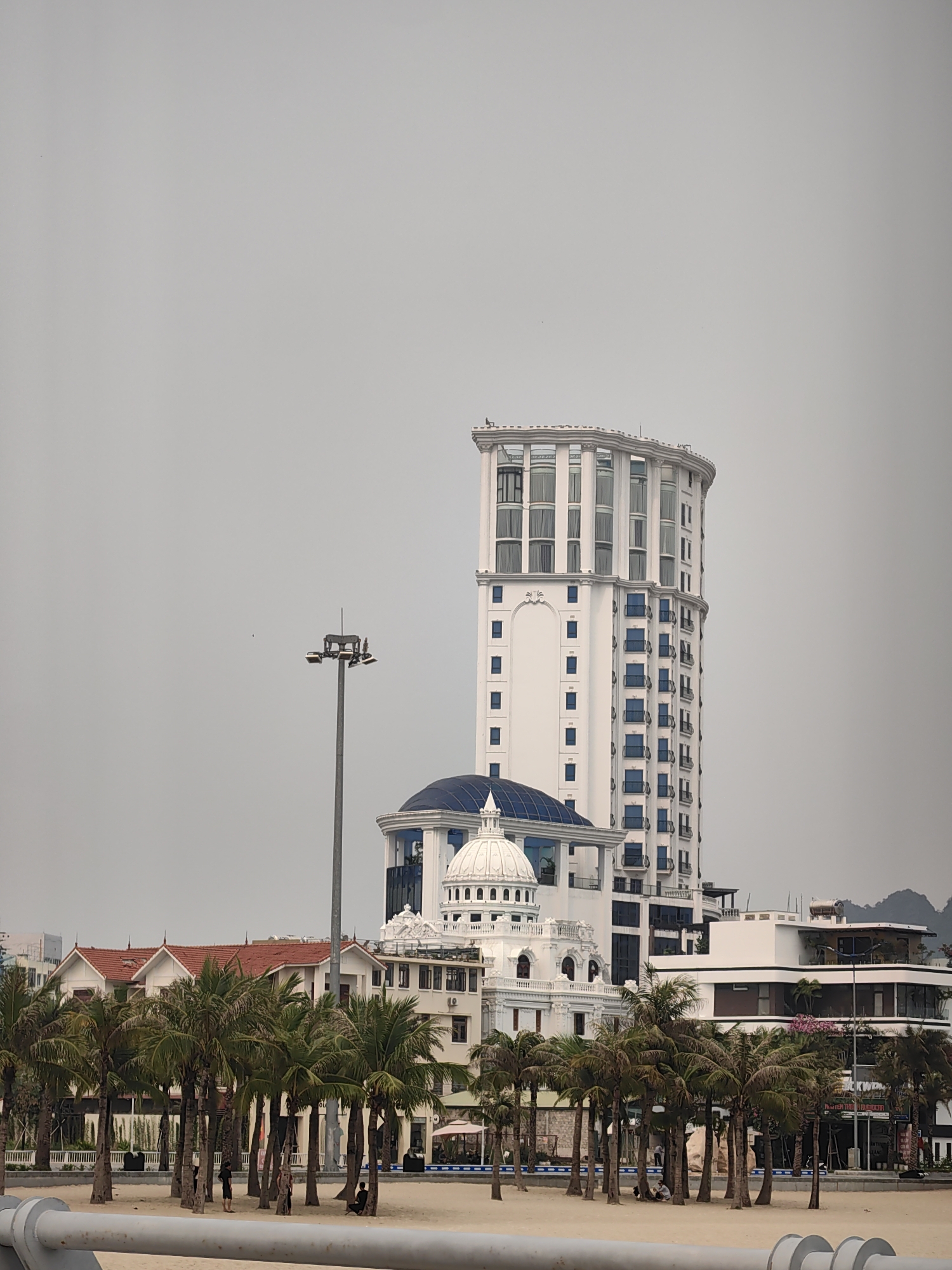 This vertical image captures a serene coastal scene in Ha Long Ward, Vietnam, under an overcast sky. In the foreground, a stretch of light-colored sand is dotted with numerous tall palm trees. Several small figures of people are visible among the trees, some standing and others appearing to be seated or resting on the sand. A grey metal railing runs horizontally across the very bottom of the frame.
Dominating the midground and background are various buildings. A tall, modern white building with a distinctive rounded top section and prominent white columns stands prominently on the right. Its upper floors feature large windows, while its lower sections have smaller blue-framed windows and balconies. Below this skyscraper, a complex of white buildings showcases classical architectural elements, including a large blue dome and a smaller white dome, along with balustrades and ornate detailing. To the left of these structures, a tall grey light pole with multiple floodlights rises into the sky. Further to the right, a contemporary white building with brown trim and a flat roof is partially visible behind the palm trees. On the far left, red-roofed houses are nestled behind more palm trees.
The sky is a uniform, pale grey, indicating an overcast day with no direct sunlight. On a sign located on the white building with brown accents on the right side of the image, the word "BLACKWOOD" is clearly visible. The overall scene suggests a quiet, possibly resort-like, beach environment.