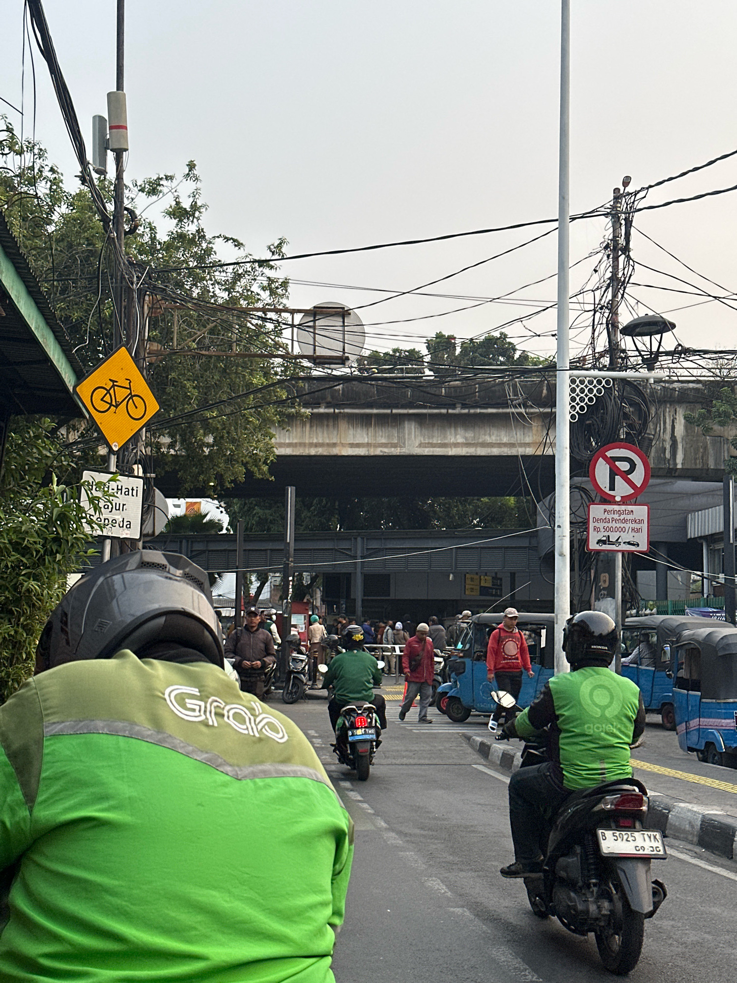 The image depicts a street scene in South Jakarta, Indonesia, with an overhead pedestrian bridge and numerous electrical wires creating a complex web above the road. The foreground is dominated by a person on a motorcycle wearing a bright green Grab jacket, with their back to the camera. Further down the road, other motorcycles and blue trishaws (bajaj) are visible, with people standing or riding them.
Several signs are present: a yellow diamond-shaped sign with a bicycle symbol, a white rectangular sign with text in Indonesian reading "Hati-Hati Laju Sepeda" (Be careful of fast-moving bicycles), and a red circular sign with a diagonal bar through a "P" symbol, indicating no parking. Below the no parking sign, another sign reads "Peringatan Denda Penderakakan Rp. 500.000/Hari" (Warning Parking Fine Rp. 500,000/Day), accompanied by pictograms of a truck and a car.
The environment suggests a bustling urban setting with typical Indonesian street transportation. The sky is overcast, indicating potentially humid weather. The overall impression is of a busy traffic area with clear regulatory signage.