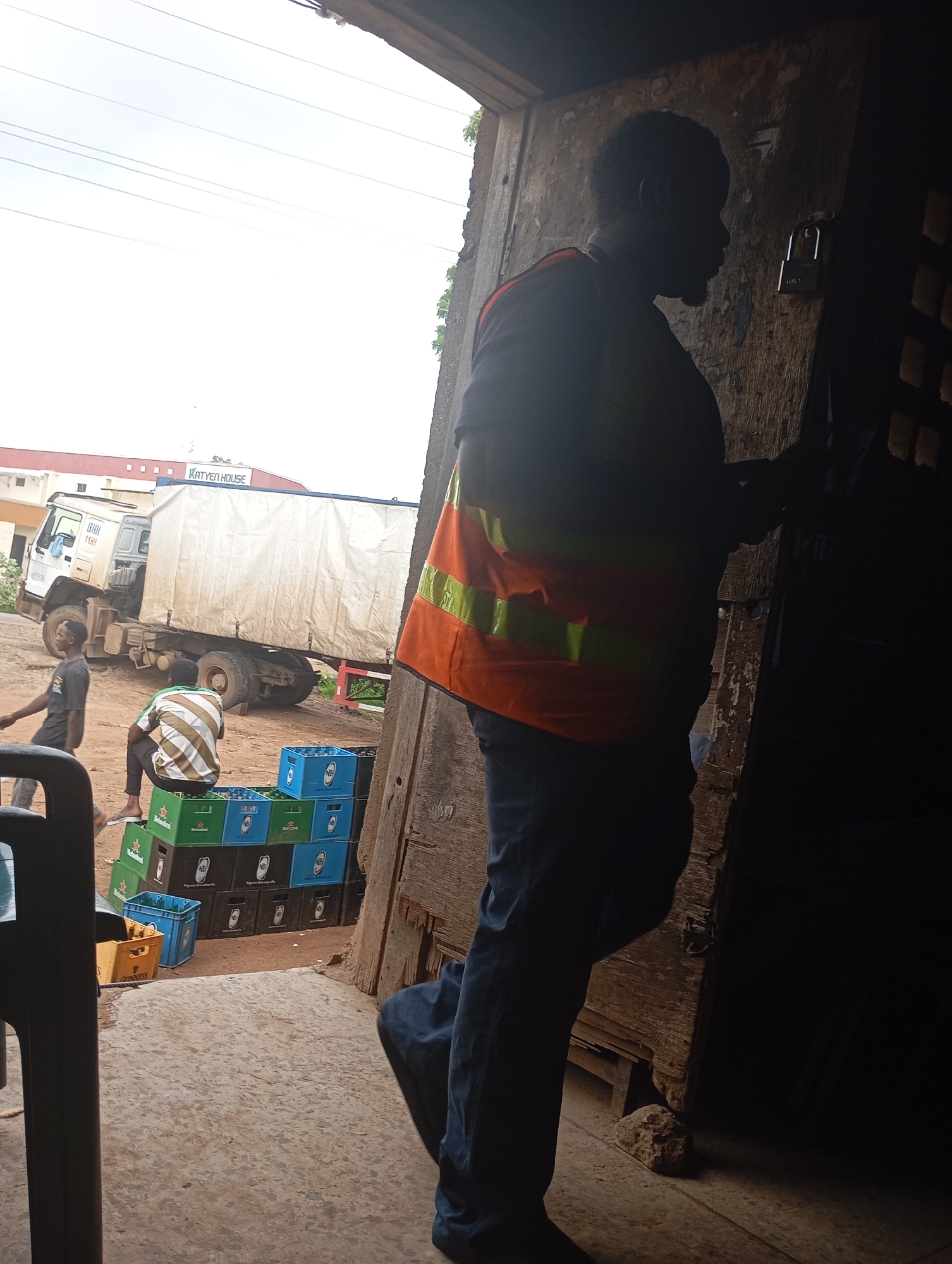 The image depicts a man wearing a reflective vest and blue jeans, standing in a doorway. Behind him, a large truck with a white tarpaulin-covered cargo area is parked. In the foreground, stacks of plastic crates, some filled with bottles, are visible. The scene appears to be an outdoor storage or loading area. The location is identified as Shendam, Nigeria.