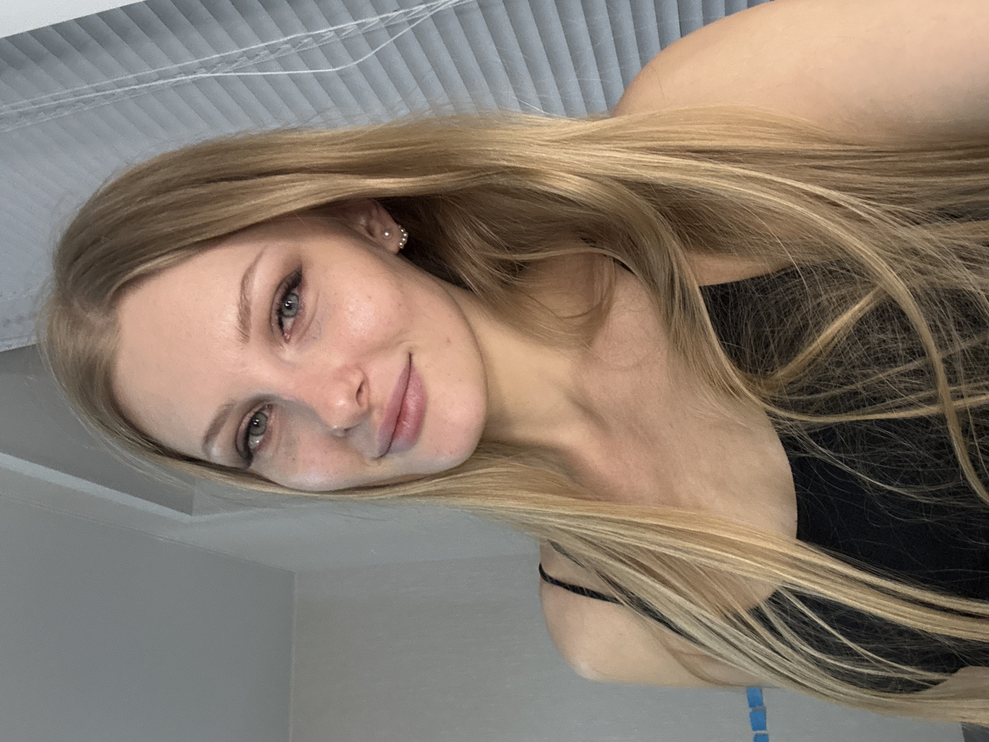 A young woman with long, blonde hair is positioned with her head tilted to the left. Her expression is subtle with a slight smile. She has fair skin and her eyes appear to be green. She is wearing a black top with thin straps. The background consists of a wall and a slatted ceiling, both in muted tones. The location in the city of Perm, Russia is not determinable from the image.