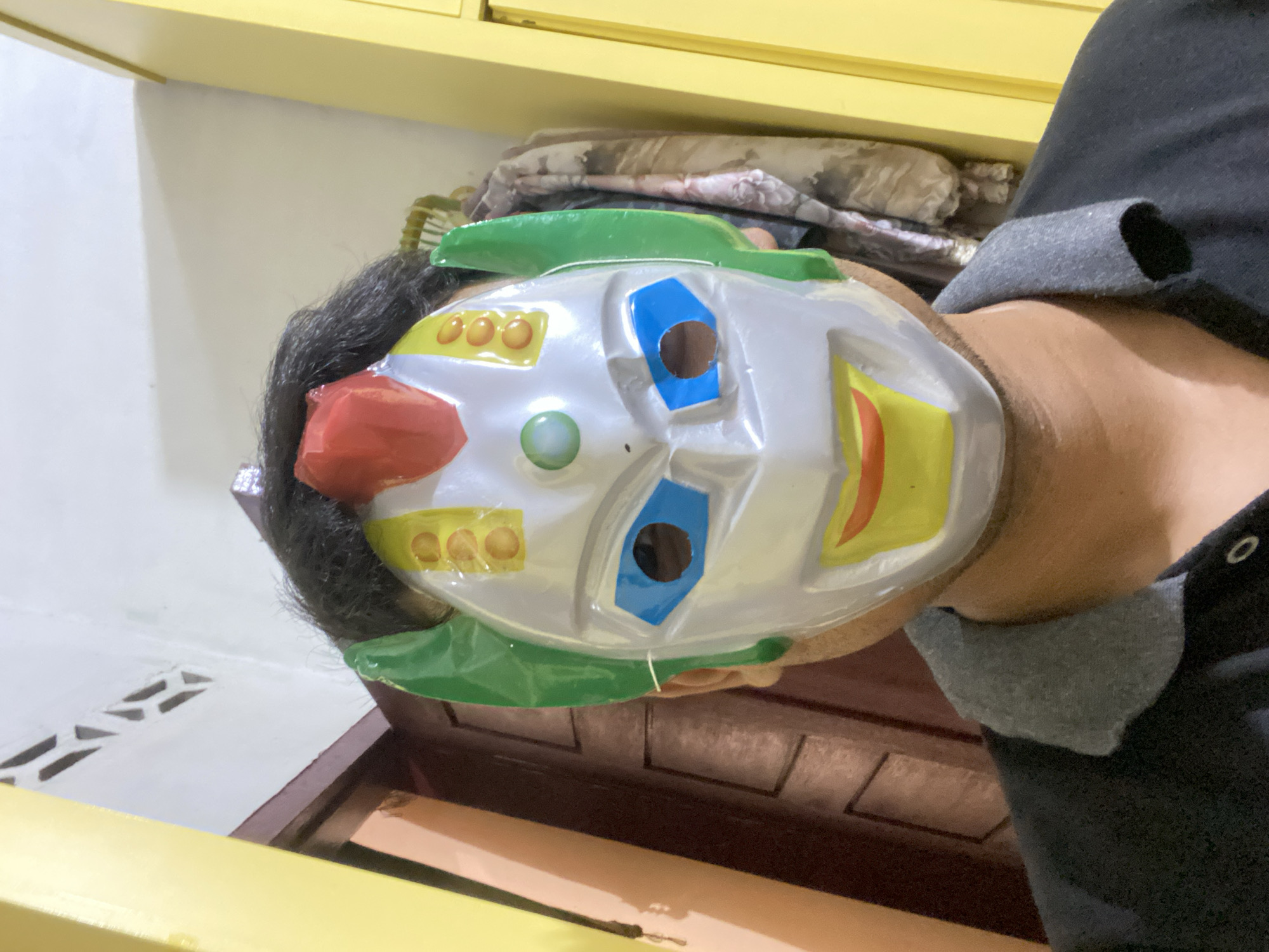 A person wearing a colorful, toy robot mask is shown from the chest up, facing the camera. The mask has a white, silver, and yellow design with blue eyeholes, green ear-like attachments, and a red crest. The person is wearing a black shirt with a grey collar. The background includes a wooden door and a yellow wall, possibly inside a room.