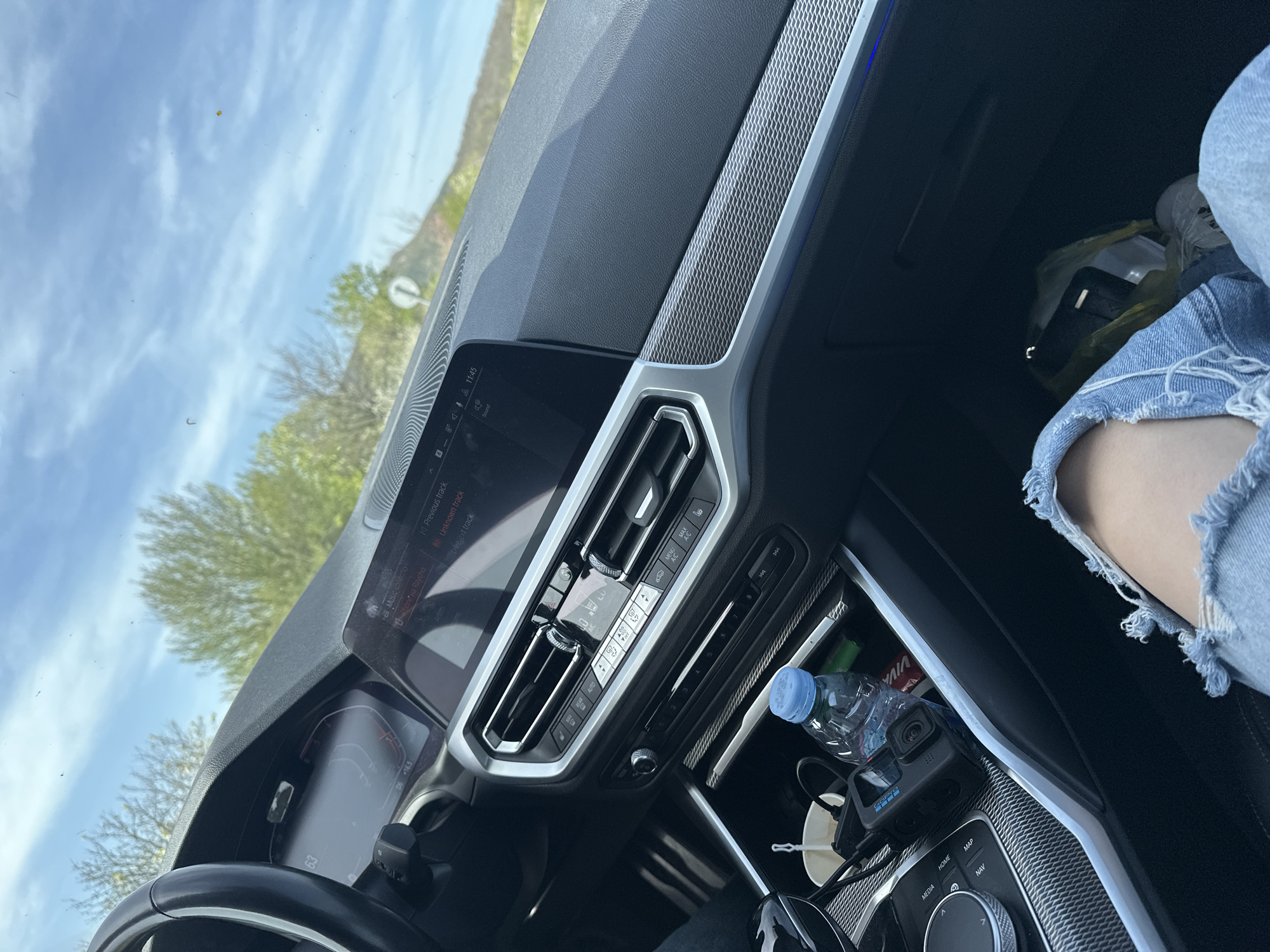 This media file captures the interior of a modern car, likely a BMW, from a passenger's perspective, looking towards the dashboard and out the front windshield. The scene appears to be set in Madrets, Bulgaria, during a bright, clear day with blue skies and scattered white clouds.
On the right side of the image, a person's leg, clad in distressed light-wash denim jeans, is visible, suggesting a relaxed passenger. The car's dashboard features a prominent infotainment screen displaying "11:45", indicating late morning. The screen also shows "Music collection", "Phone", "on Raycho", "Previous track", "Unknown track", "Delete track", "Sound", and "Artists". Below the screen, the climate control panel includes buttons for "AUTO", "MAX A/C", fan speed, temperature, and seat heating.
The central console houses an iDrive controller with surrounding buttons labeled "MENU", "MEDIA", "HOME", "MAP", "NAV", "BACK", and "OPTION". Visible in the storage compartment near the gear shifter are a blue-capped water bottle, a black GoPro camera (GoPro Hero 9 Black, recognizable by its design and branding), and a small white cup. A red "VIVA" snack wrapper is also visible. In the footwell or glove compartment area, a yellow plastic bag containing a white styrofoam container and a dark-colored object (possibly a phone or wallet) is present.
Through the windshield, green trees with spring foliage and a partial view of a road sign (a circular sign with a diagonal line) are visible. The driver's instrument cluster shows "63" (likely speed) and "D" (drive mode), along with "-16.5", which could be an outside temperature reading. The overall impression is of a casual car journey under good weather conditions.