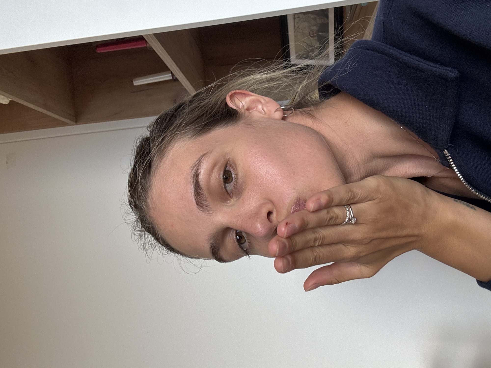 A close-up, indoor photograph shows a woman with fair skin, light brown hair, and brown eyes covering her mouth with her right hand. She is wearing a navy blue zippered jacket and a silver ring on her ring finger. Her right ear is visible, adorned with a small silver hoop earring. The background consists of a white wall and what appears to be a wooden shelf or cabinet. The location is Unknown, Unknown.