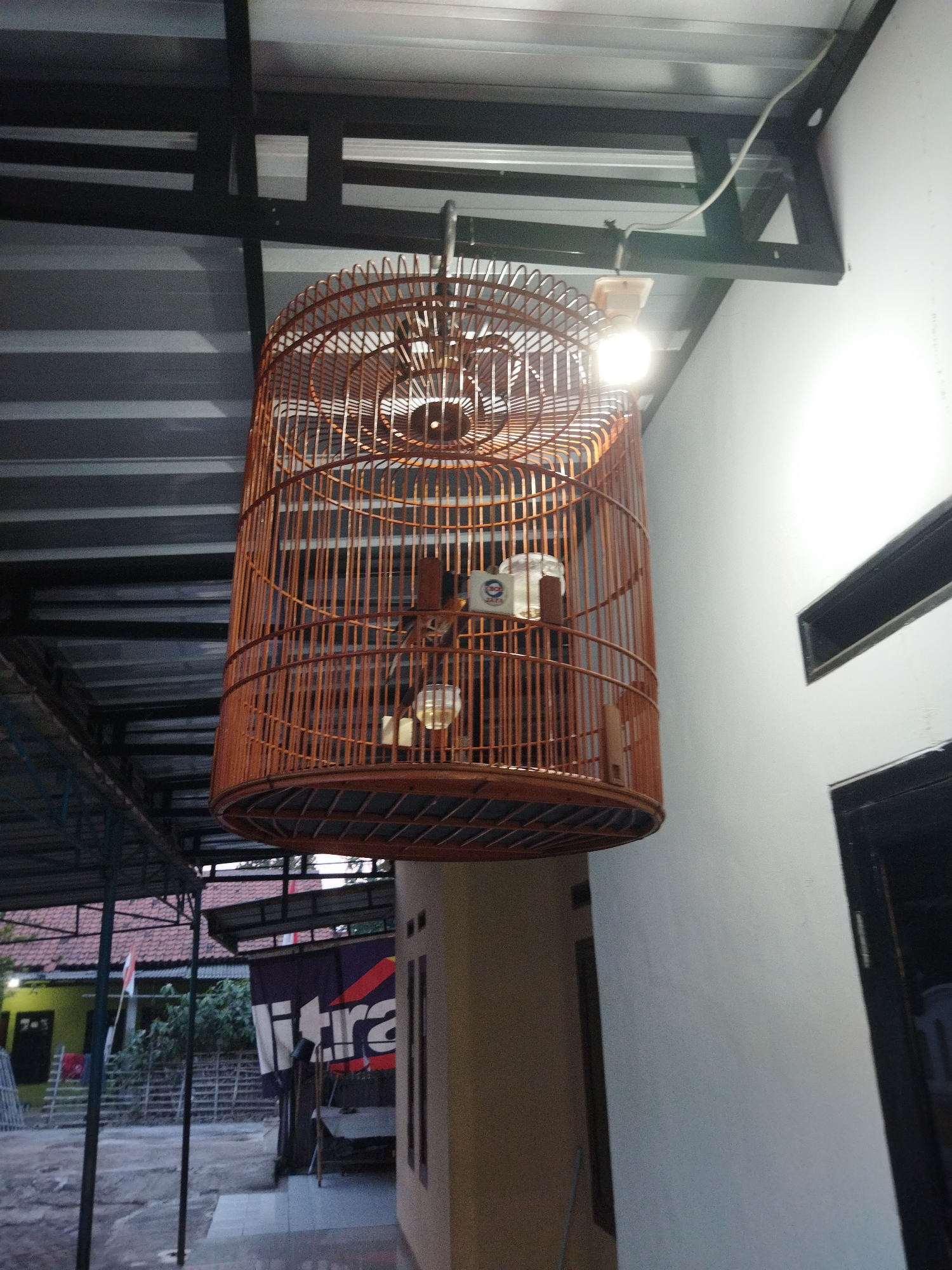 In Kosambi, Indonesia, a bamboo bird cage is suspended from an outdoor ceiling structure, illuminated by an overhead light fixture. The cage appears to be empty. Below and to the left of the cage, a building with a corrugated metal roof is visible, along with a fence and some vegetation. To the right, a white wall with dark-framed windows forms part of a building. The scene is outdoors, under a covered area, suggesting a residential or commercial setting in the urban environment.