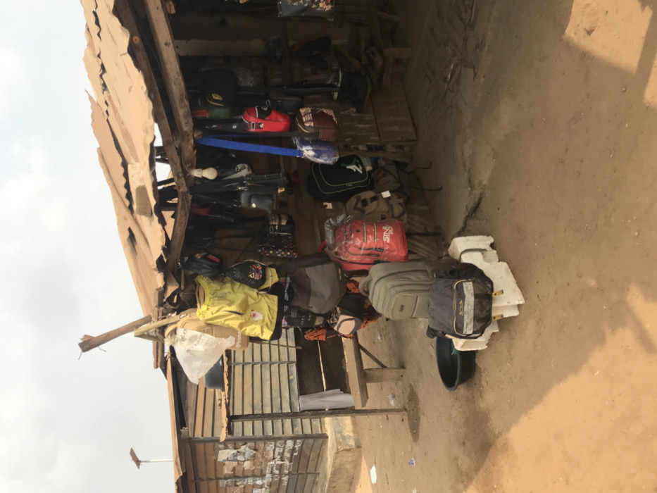 The image displays an outdoor market stall located in Ogbese, Nigeria. The stall features a rustic construction with rough wooden beams and posts supporting a worn, tan-colored corrugated metal roof exhibiting visible damage. Hanging from the wooden framework are numerous bags and backpacks of varying sizes and designs, including yellow, red (with "SINCE 1990" visible), grey, black, blue, and patterned items, some still in clear plastic wrapping.
On the reddish-brown, dusty ground in front of the stall, a grey backpack is positioned atop two white concrete blocks, alongside a dark blue messenger bag. A black plastic basin rests on the ground nearby. To the left of the main stall, an adjacent building with corrugated siding or horizontal wooden planks has a simple wooden bench affixed to its exterior, draped with a light-colored fabric. A single bare light bulb hangs from the interior roof structure of the stall. In the upper left background, a street light pole equipped with a solar panel is discernible against an overcast sky. No individuals are present in the scene, and no actions or interactions are depicted.