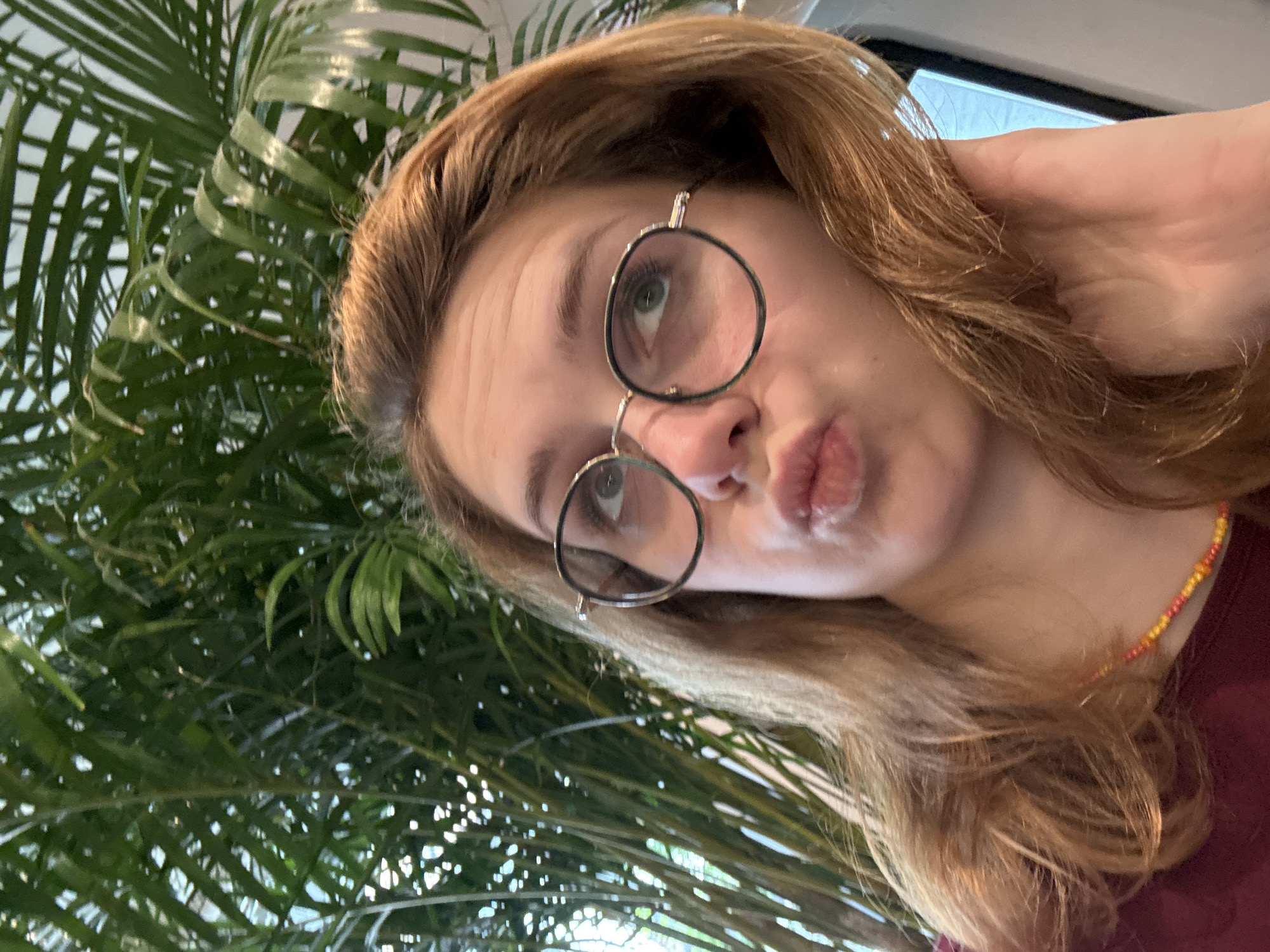 A woman is posing indoors, wearing round-frame glasses and a beaded necklace. Her lips are pursed. A large potted palm plant is visible in the background. No information about the location in the city of Kragujevac, Serbia, is discernible from the image.