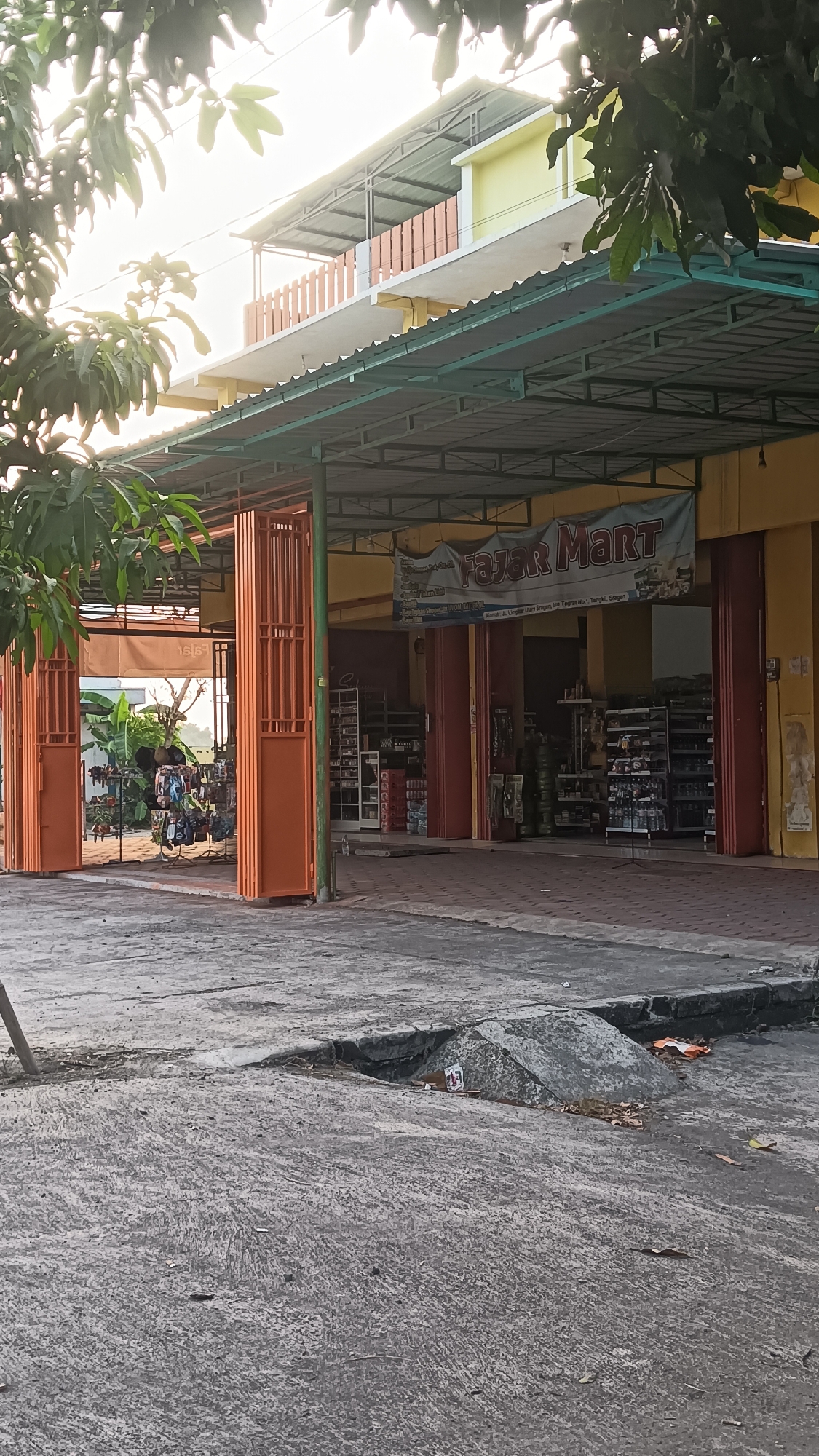 The image captures a street-level view of a commercial establishment, likely a minimarket, identified by the banner reading "FAJAR MART". The storefront is open, with orange accordion-style doors partially retracted, revealing shelves stocked with merchandise. The building appears to be two stories, with a second-floor balcony and an awning extending over the storefront. The area in front of the mart is paved, with a section of broken concrete in the foreground. Trees with lush green foliage overhang the scene, and power lines are visible against the sky. The lighting suggests it is either early morning or late afternoon. The location is specified as Karang Tengah, Indonesia.