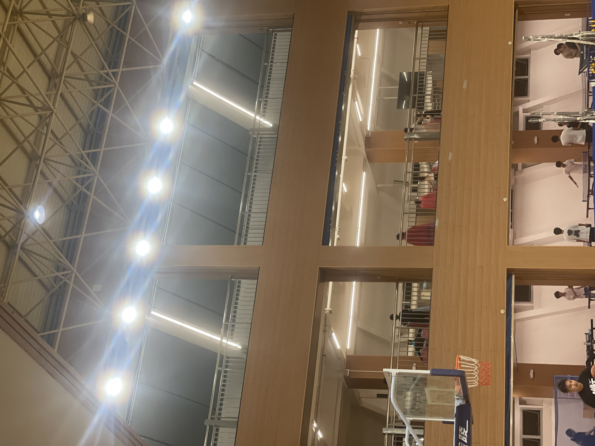 An indoor gymnasium or sports hall is captured from a low, upward-looking angle, revealing a multi-level modern facility in Chongqing High-tech Zone, China. The ceiling features a complex metallic truss structure with numerous bright, circular spotlights casting strong light and lens flares.
On the right side of the image, multiple vertical sections of light-colored wooden paneling divide views into various levels of what appear to be viewing balconies or upper rooms. These levels are illuminated by linear strip lights and equipped with metal railings. Several individuals are visible on these upper levels, some standing and some seemingly observing activity below. They appear to be young adults or teenagers.
In the foreground, partially visible on the right, is a basketball hoop with an orange net attached to a clear backboard and a blue support structure. Chinese characters are visible on the blue support, likely indicating a brand or facility name. Below the hoop, on the main court level, a person in dark attire, possibly sportswear, is visible, suggesting active participation or spectating. Other figures are also faintly seen in the distance on the court level.
The facility showcases contemporary architecture with a combination of wood paneling, glass or reflective surfaces, and metal accents. The dominant artificial lighting suggests the photograph was taken indoors, possibly in the evening or at night. The overall impression is that of a bustling recreational or sports event taking place within a well-lit, modern setting.