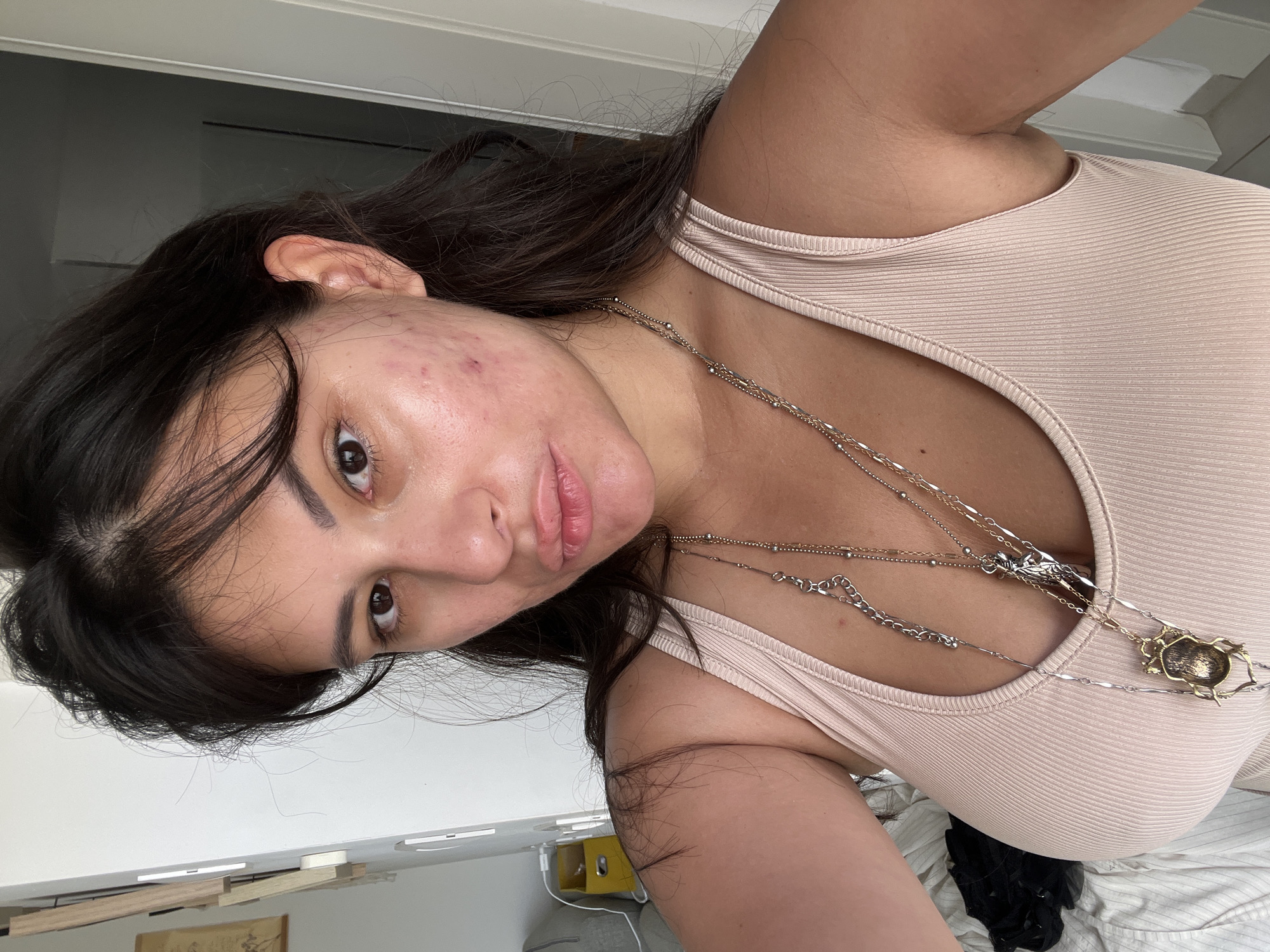 A young woman with dark hair and tanned skin, exhibiting acne on her face, is pictured in a close-up, selfie-style shot. She is wearing a light beige, ribbed tank top adorned with multiple layered necklaces, one featuring a beetle pendant. The background appears to be an indoor setting, possibly a bedroom, with white walls, a partially visible wooden structure on the left, and a light-colored door on the right. The lighting suggests natural daylight. The location is identified as Kiryat Bialik, Israel.
