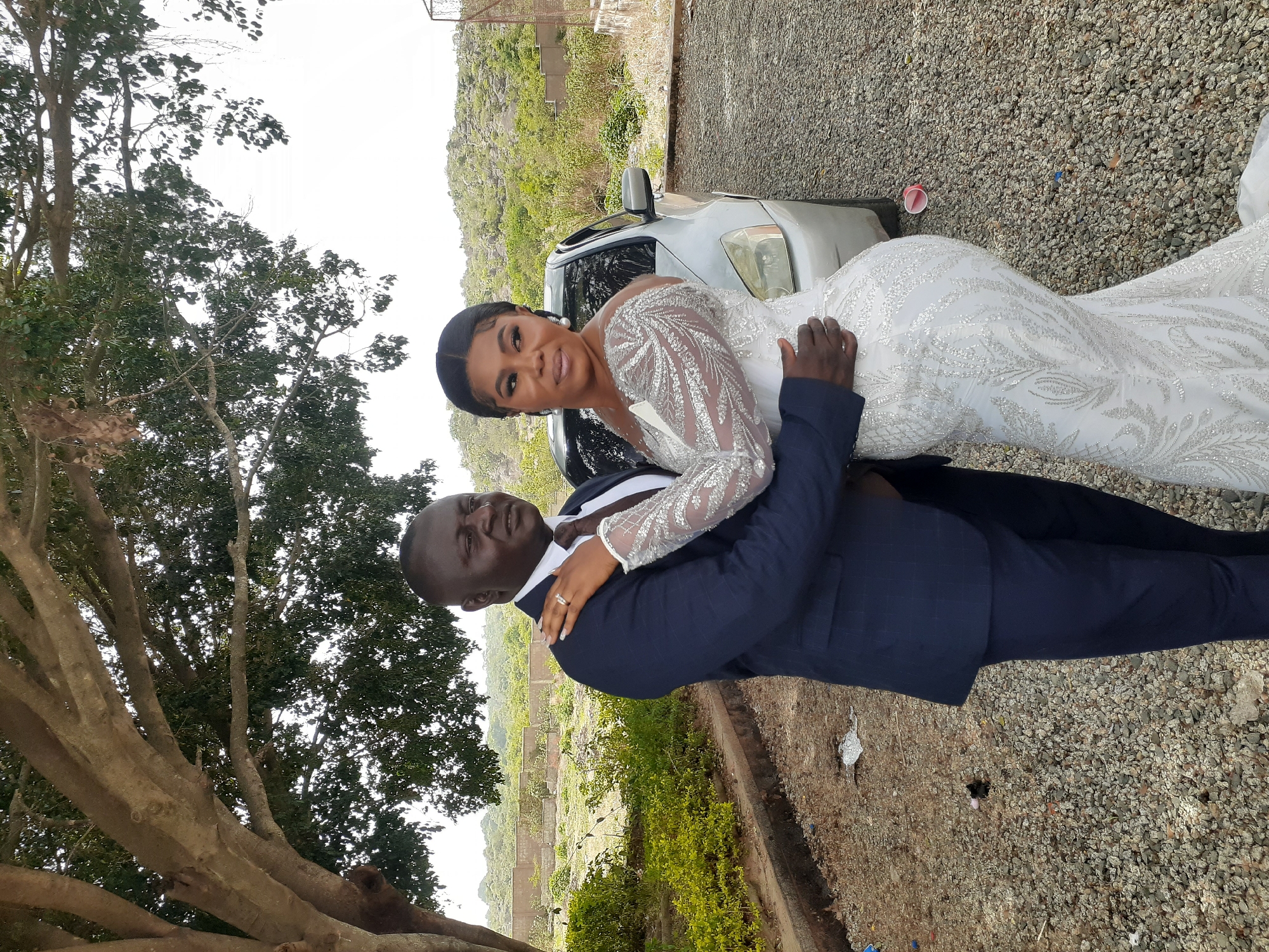 In Jos, Nigeria, a bride in a white, long-sleeved, embellished gown is being held in the arms of a groom wearing a navy blue suit. They are positioned on a gravel surface next to a silver car. The background features a lush landscape of green vegetation and trees, and a mountain is visible in the distance. A large tree is on the left side of the image.