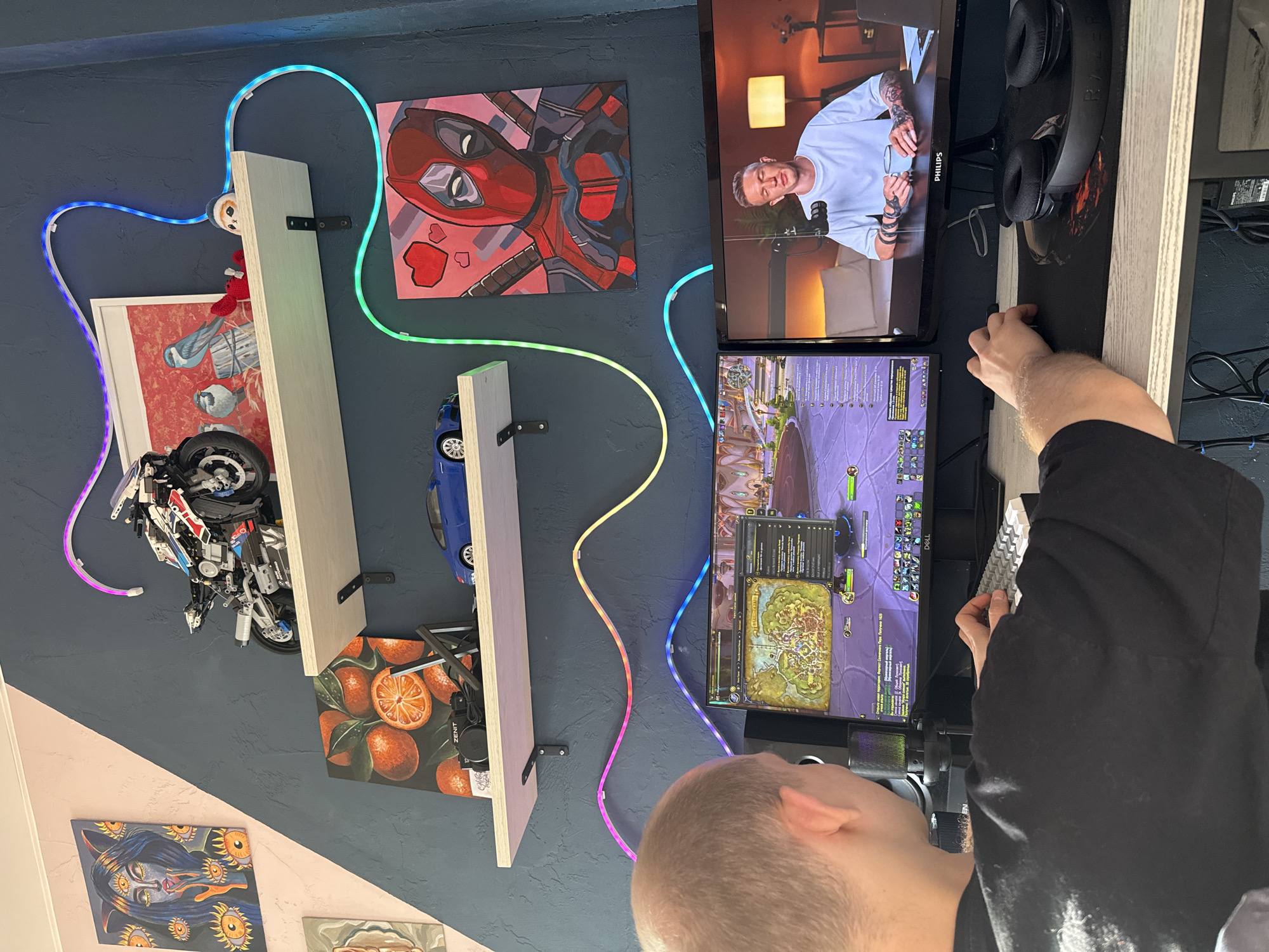 A person is playing the video game World of Warcraft on a dual monitor computer setup. The top monitor displays a video of a man in a white shirt. The person is using a white keyboard and a black mousepad with a Razer headset on it. Behind the computer are three shelves with various objects on them, including a Lego motorcycle, a blue car, a painting of oranges, and a Deadpool painting. A color-changing LED strip is running along the wall behind the shelves. There is a painting of a cat-like creature on the wall. The location is likely indoors, but the city of Rostov-on-Don, Russia is not determinable from the image.