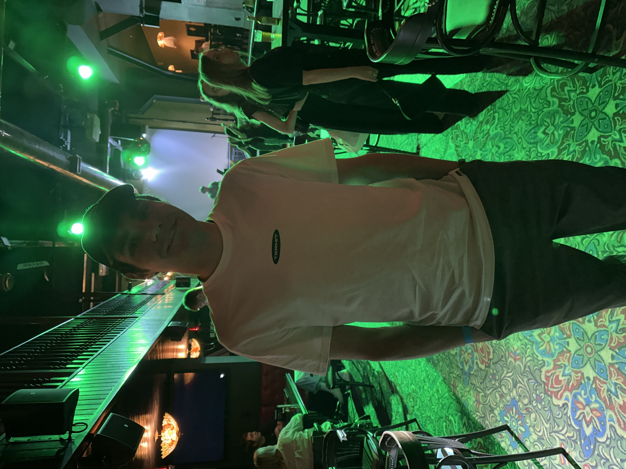 The image captures a medium shot of a man standing indoors, facing the camera. He is wearing a white t-shirt with a green logo and dark pants, topped with a dark cap. His hands are in his pockets. The man is positioned in the foreground, with the background showing a dimly lit interior. Green lighting illuminates the scene, highlighting a balcony-like structure above and various figures in the mid-ground. On the balcony, there appears to be a railing and some equipment. In the mid-ground, several individuals are visible, some seated and others standing. A stage with a projection screen is also discernible in the distance. The floor is covered with a patterned carpet. The presence of a stage and audience seating suggests the location could be a bar, club, or music venue. There is no visible information in the image to suggest the city of Saratov, Russia.