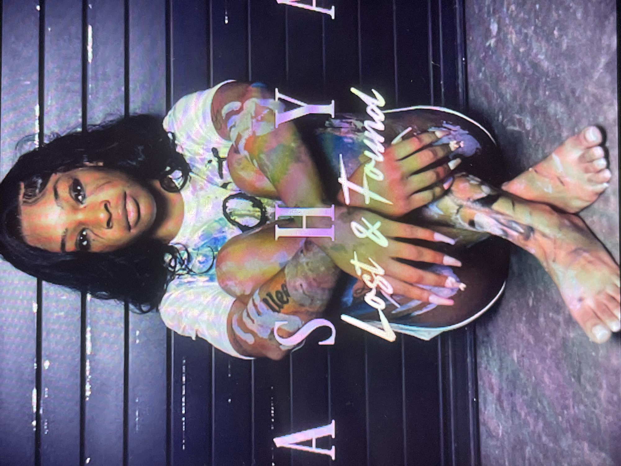 This media file is a portrait of a young woman taken from an overhead perspective, presented in a vertically oriented shot. The subject, a woman with dark hair and a dark complexion, is positioned lying down with her knees drawn up towards her chest, her arms wrapped around her legs. She is wearing a light-colored, possibly tie-dye, t-shirt and dark bottoms. Her skin, particularly on her arms, legs, and face, as well as her clothing, is streaked with various colors of paint, suggesting an artistic or performance-related context. Her long fingernails are painted a light, possibly lavender, color. A tattoo with what appears to be the word "Never" is partially visible on her forearm.
The woman is looking directly at the camera with a neutral expression. She is set against a dark, ribbed background that resembles vertical panels or slats, possibly an indoor studio setting. The overall lighting is dim, but the colors on her body are vibrant.
Several large, stylized words are overlaid on the image. The word "SHY" is prominently displayed vertically in light-colored, shadowed letters, running down the center-right of the frame. The phrase "Lost & Found" is written horizontally in a flowing script, positioned on the right side of the woman's body. A partial letter "A" in a similar style is visible in the bottom-left corner.
The image captures a posed moment, emphasizing artistic expression through body paint and overlaid text. The setting appears to be a controlled environment, likely for a photoshoot. This media file is presented with the contextual location of Sofia, Bulgaria.