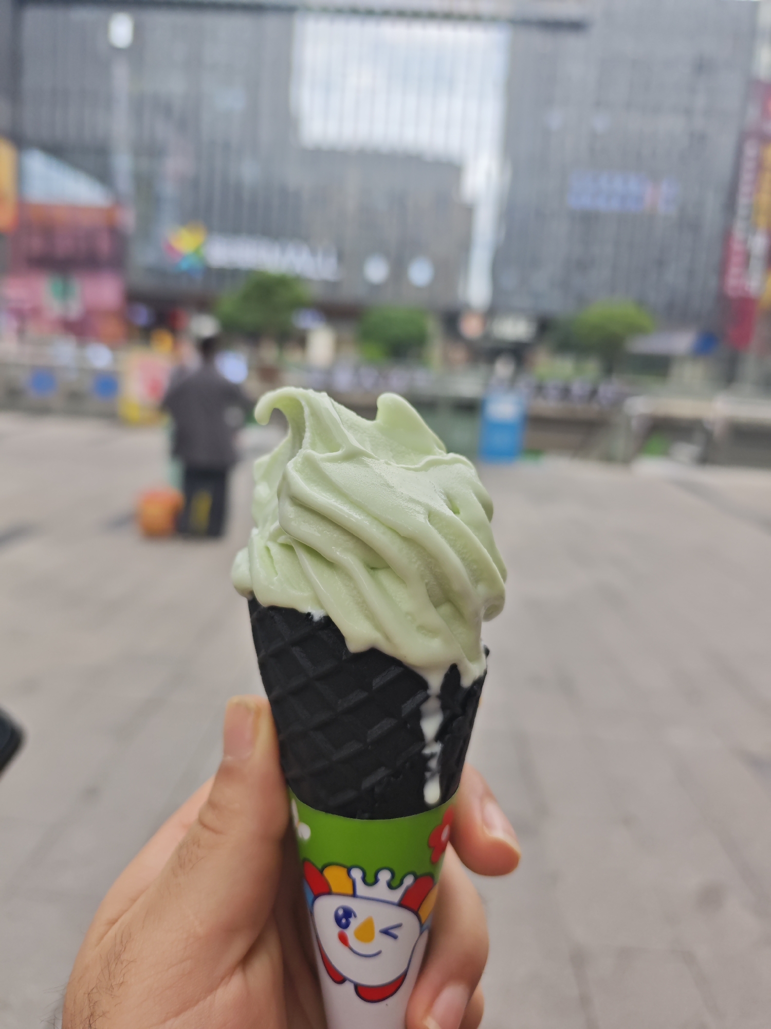 A person is holding a green ice cream cone with a black waffle cone and a cartoon character on the wrapper. The ice cream is slightly melted. In the background, there is a blurred cityscape, a pedestrian walking with luggage, and a large building that says "ARRIVAL HALL". The location is in the city of Chongqing High-tech Zone, China.