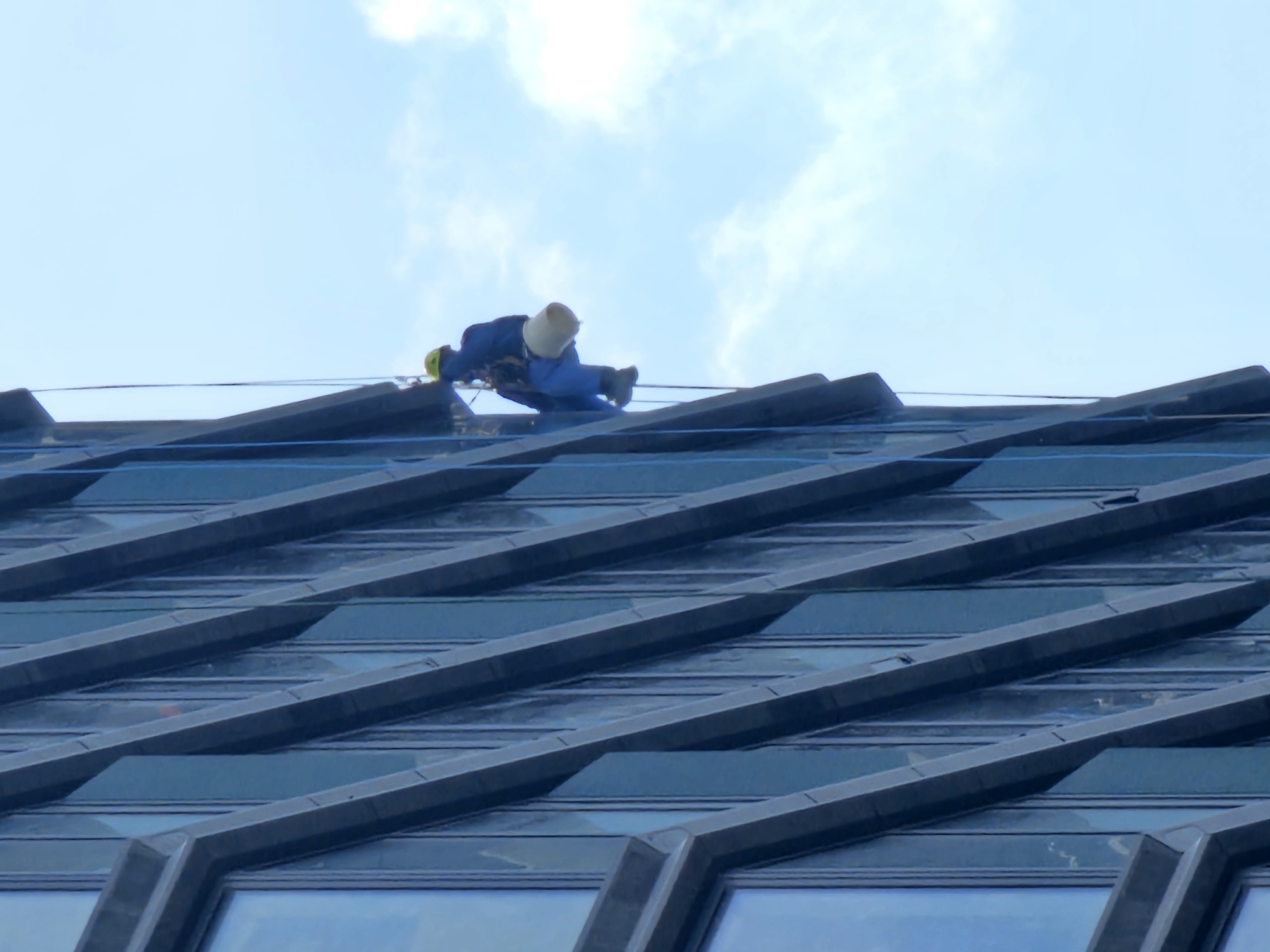 A worker in blue overalls, a yellow hard hat, and a beige bucket hat is secured with safety gear on the slanted roof of a building. The building features a patterned facade of angled metal beams and what appear to be large windows, giving it a modern architectural design. The sky in the background is light blue with some wispy white clouds. The location is Moscow, Russia.