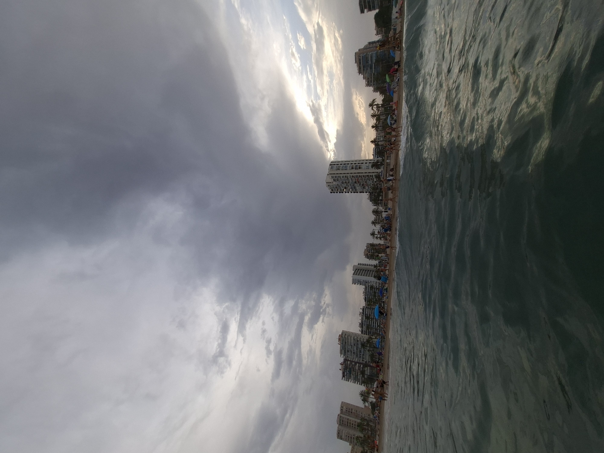 The image shows a beach scene in Alicante, Spain. In the foreground, the ocean water is visible with small waves. The middle ground features a sandy beach with people scattered across it, some walking, others relaxing on lounge chairs. Several beach umbrellas are set up, offering shade. In the background, a row of modern, multi-story apartment buildings lines the coast. The sky is partly cloudy, with dramatic, dark clouds dominating the upper portion of the frame, contrasting with patches of lighter sky and the glow of the setting sun peeking through. Palm trees are interspersed among the buildings, indicating a warm climate.