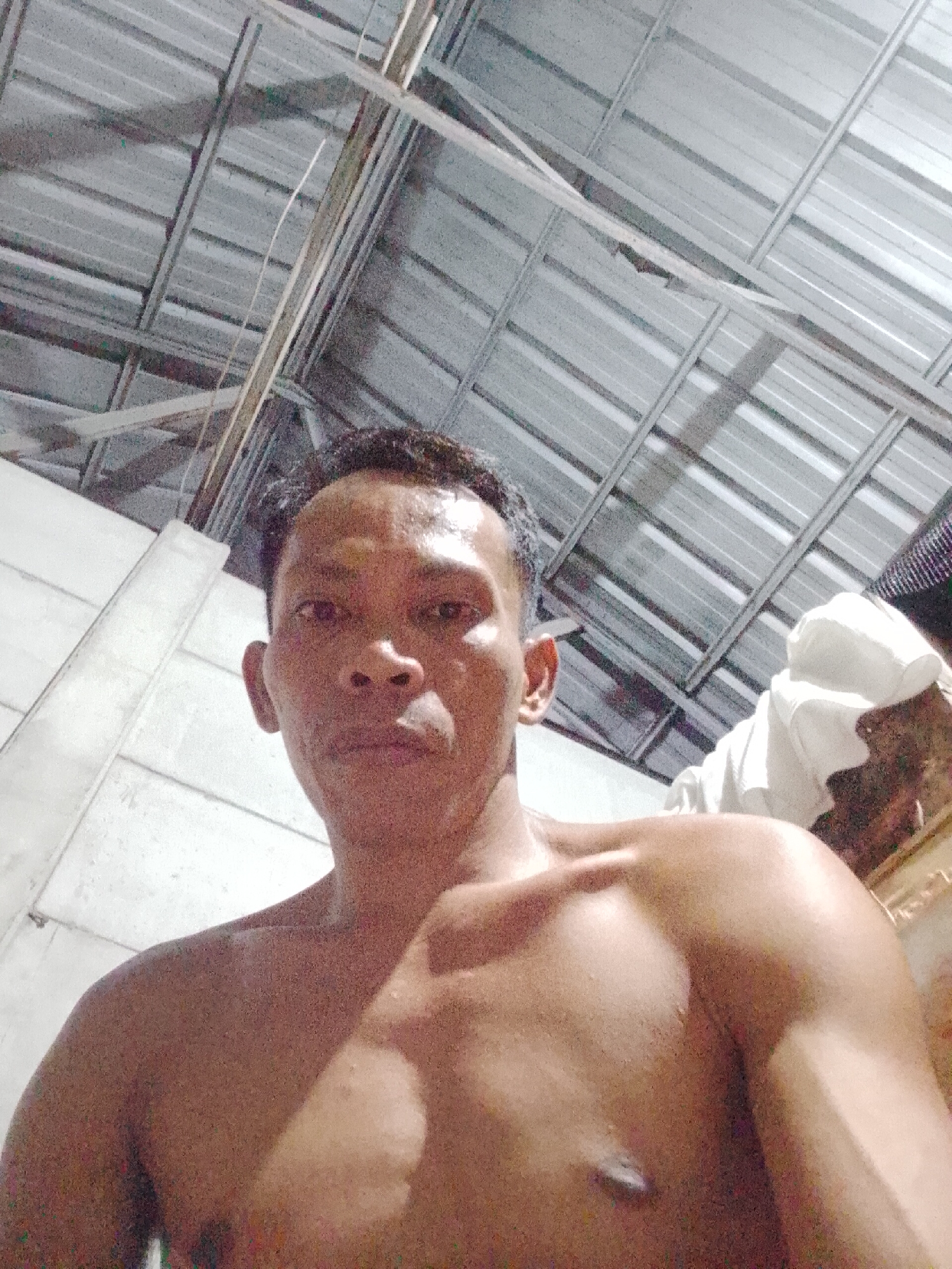 A shirtless man is positioned in the foreground, facing the camera. He appears to be in an interior space, indicated by the corrugated metal ceiling and white wall in the background. A white object, potentially a cloth or garment, is partially visible on the right side of the frame. The image seems to have been taken from a low angle. The city of Bukit Ketok, Indonesia is presumed to be the location based on the prompt.