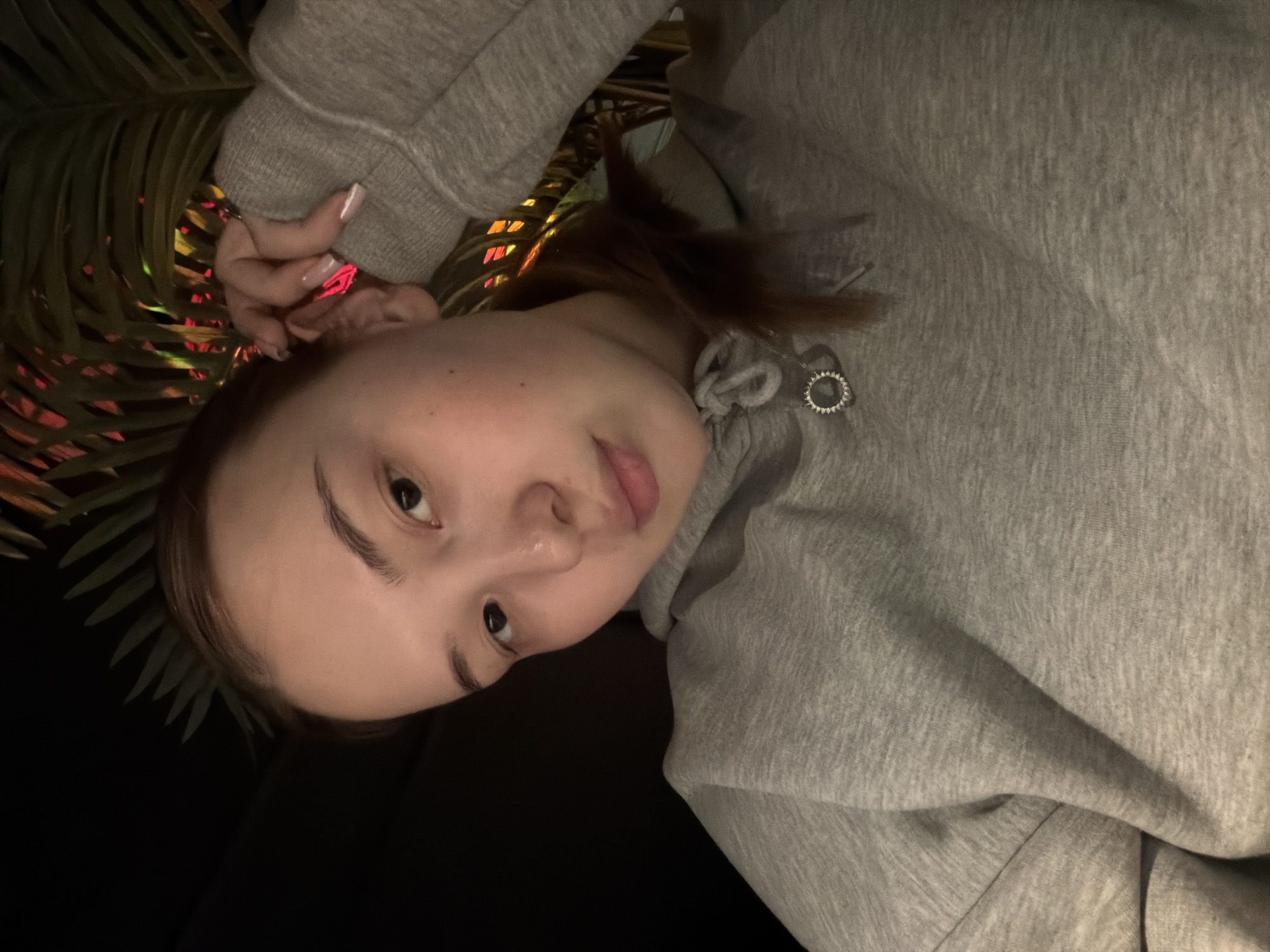 A woman with fair skin and brown hair is wearing a gray hooded sweatshirt. She is holding her hand near her ear. In the background is a palm leaf illuminated with red and yellow lights. The image appears to be taken indoors in low light. The location is possibly Chaikovsky, Russia.