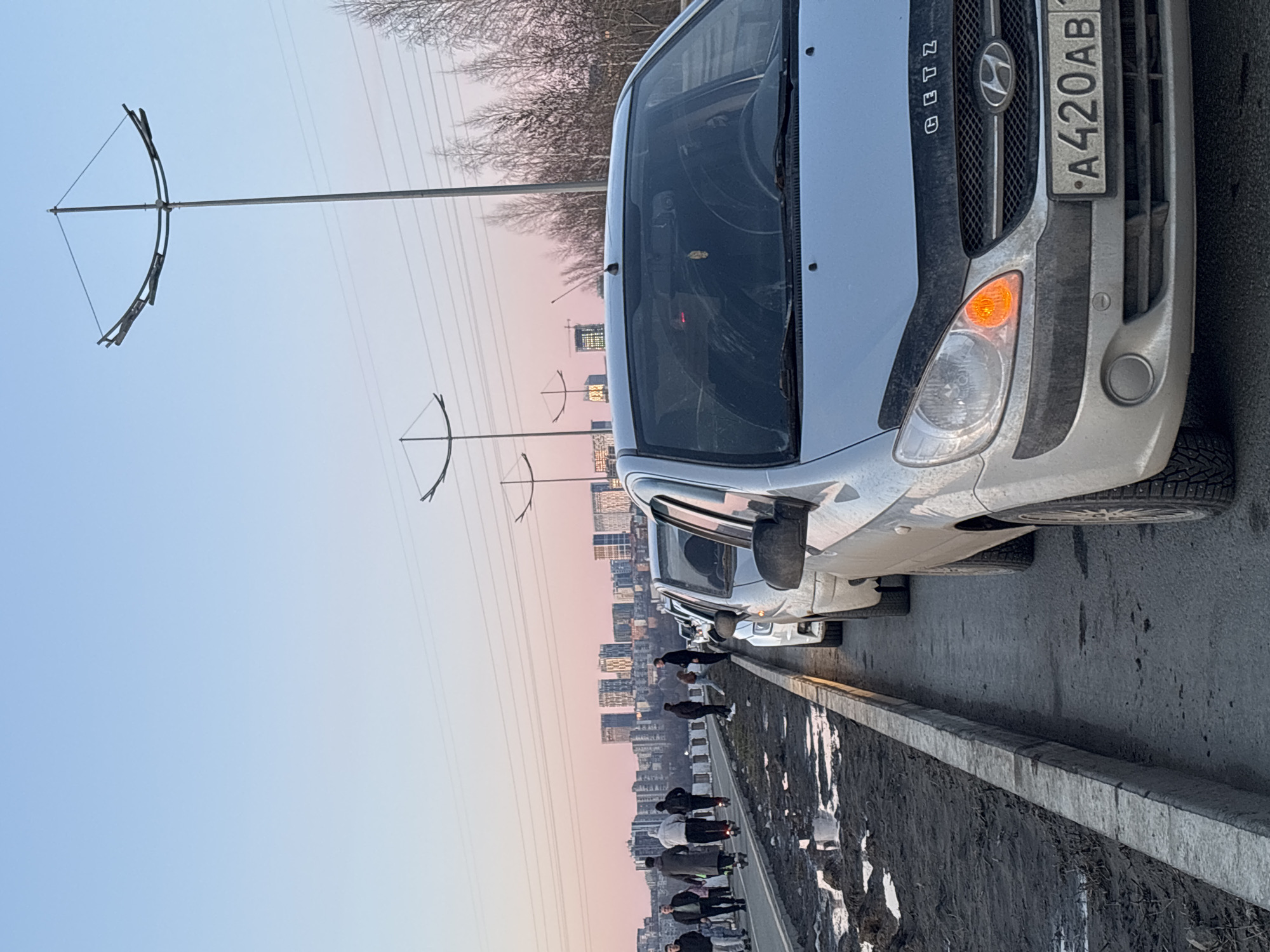 The image shows a row of parked cars on a road in Izhevsk, Russia. A Hyundai Getz is in the foreground. A line of pedestrians walks along the edge of the road, near a curb. The backdrop features a cityscape with tall buildings under a sky transitioning from blue to orange at sunset. Unique streetlights are visible along the road. The license plate of the front car reads "A420AB" and appears to be a Russian license plate.