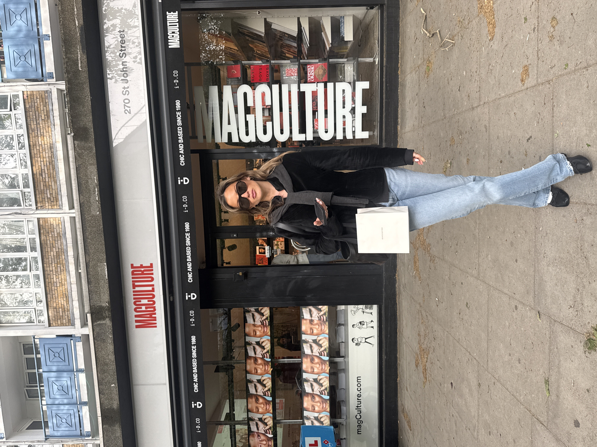 An eye-level shot captures a woman standing on a sidewalk outside a shop called "MAGCULTURE" on St John Street in London, United Kingdom. The image is rotated 90 degrees clockwise, placing the woman horizontally in the frame.
The woman is positioned centrally, dressed in a black long-sleeved top or jacket, light blue jeans, and black shoes. She wears dark sunglasses and has light brown hair cascading over her shoulders. In her left hand, she holds a white shopping bag, while her right hand holds a mobile phone. Her posture is relaxed, with her legs slightly crossed at the ankles.
The shopfront behind her features large glass windows displaying various magazines and books. The prominent shop name "MAGCULTURE" is visible in large white letters across the main window, and in smaller red text on an awning above. Below the main sign, text repeatedly states "CHIC AND BASED SINCE 1980" and "i-D.CO". A website address, "magCulture.com", is also visible on the lower part of the window display. Inside, magazine covers show titles like "BOYS BOYS BOYS!" and "BRISHE". On the facade to the left of the shop, the address "270 St John Street" is clearly legible.
The setting appears to be a street in a city, with a concrete sidewalk. In the background, other buildings with windows and blue decorative panels on balconies are visible. The lighting suggests it is daytime, likely overcast, with soft shadows. The overall scene depicts a casual urban moment, possibly after a shopping trip.