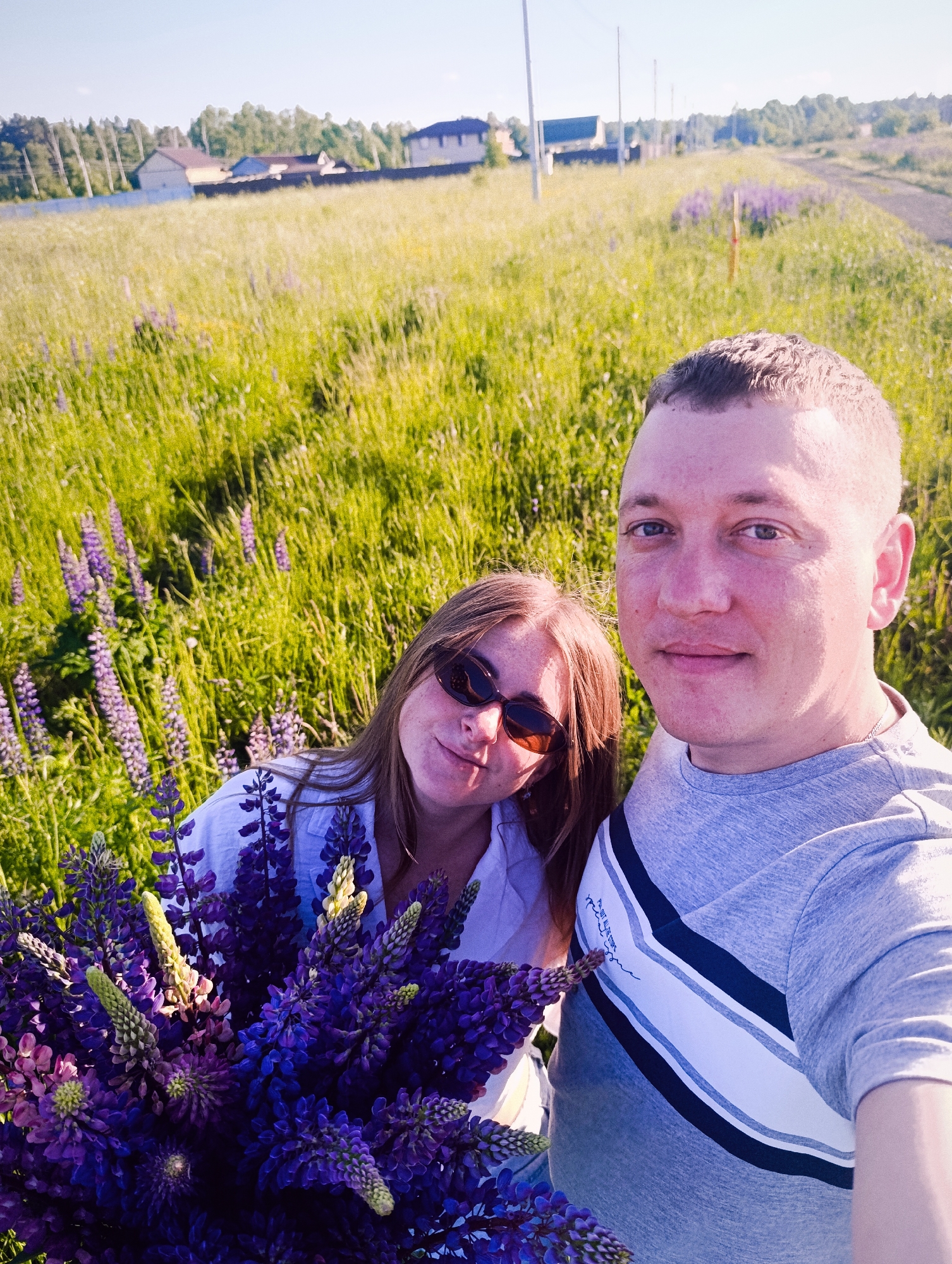 A selfie-style outdoor photograph depicts an adult male and female in a rural or suburban landscape under a clear sky. The male, positioned on the right, has short hair, a slightly reddish complexion, and wears a grey t-shirt with a white and dark blue chevron design. He faces the camera directly. The female, to his left, has long reddish-brown hair, wears sunglasses, and a light-colored collared shirt, holding a substantial bouquet of purple lupine flowers. She smiles faintly. The immediate surroundings consist of a field of green grass interspersed with numerous purple lupine flowers. In the mid-ground and background, multiple houses, a line of trees, and a dirt path with utility poles are visible. Location: Unknown, Unknown.