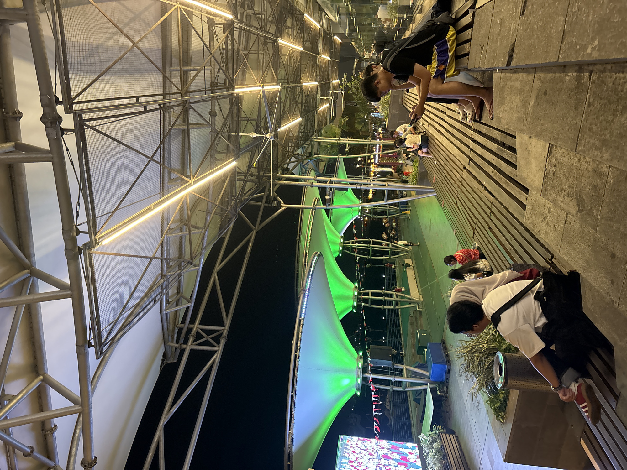 An outdoor seating area at night is depicted in North Jakarta, Indonesia, under a large, open-air canopy supported by a metal framework. The canopy features several large, cone-shaped sections illuminated with vibrant green light. Rows of fluorescent lights are visible along the underside of the metal structure, casting a warm glow.
In the foreground, a group of people are seated on tiered wooden benches and stone steps. On the right side, two young men are seated, one of whom is looking at his phone. Further to the left, other individuals are seated on the benches, some facing a large screen displaying an image of people in festive attire. Behind them, palm trees and other vegetation are visible, suggesting a tropical or subtropical environment. The overall atmosphere appears to be a public gathering space, possibly an entertainment or dining venue.