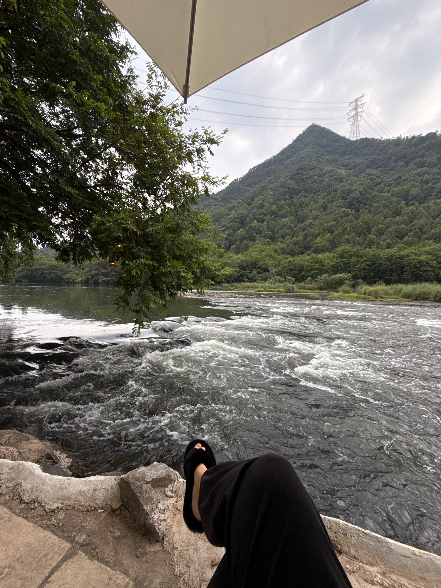 The image depicts a serene natural scene near Yiwu, China, focused on a person relaxing by a flowing river. The perspective is from someone seated on a stone embankment, with their legs and feet, clad in black trousers and fluffy black slippers, prominently in the foreground. The river itself is a central element, characterized by rapid currents and white water, suggesting a shallow, rocky riverbed. To the left, lush green trees overhang the water, their branches casting shadows. Beyond the river, a densely forested mountain range rises, and a large electrical transmission tower stands atop one of the peaks. The sky is overcast, contributing to the subdued lighting of the scene. An off-white umbrella or canopy extends from the top of the frame, providing shade. The overall impression is one of tranquil leisure in a natural setting outside the urban environment of Yiwu.