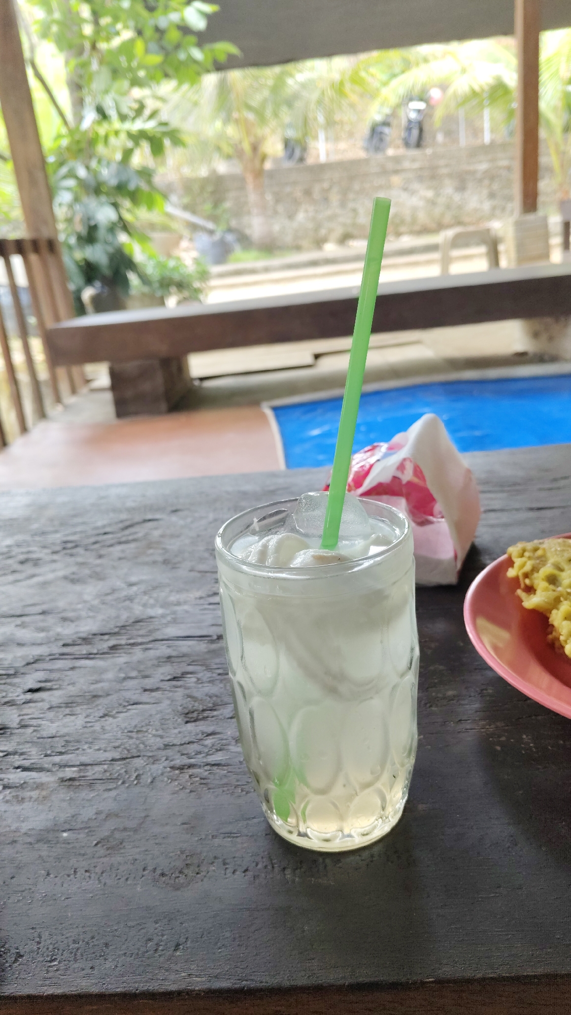 A close-up, eye-level shot focuses on a glass of white liquid with ice and a green straw, placed on a dark, weathered wooden table. To the right of the glass, a pink plate holds a portion of yellow, fried food. In the background, a red and white object, possibly packaging, is partially visible behind the glass. Beyond the table, the environment appears to be an outdoor seating area, with blurred foliage, palm trees, a stone wall, and a blue surface, possibly a pool, suggesting a tropical or semi-tropical location. The overall scene indicates a casual dining or refreshment setting. There are no people visible in the image. The image is likely taken in Kresek, Indonesia, given the presence of palm trees and the general aesthetic.