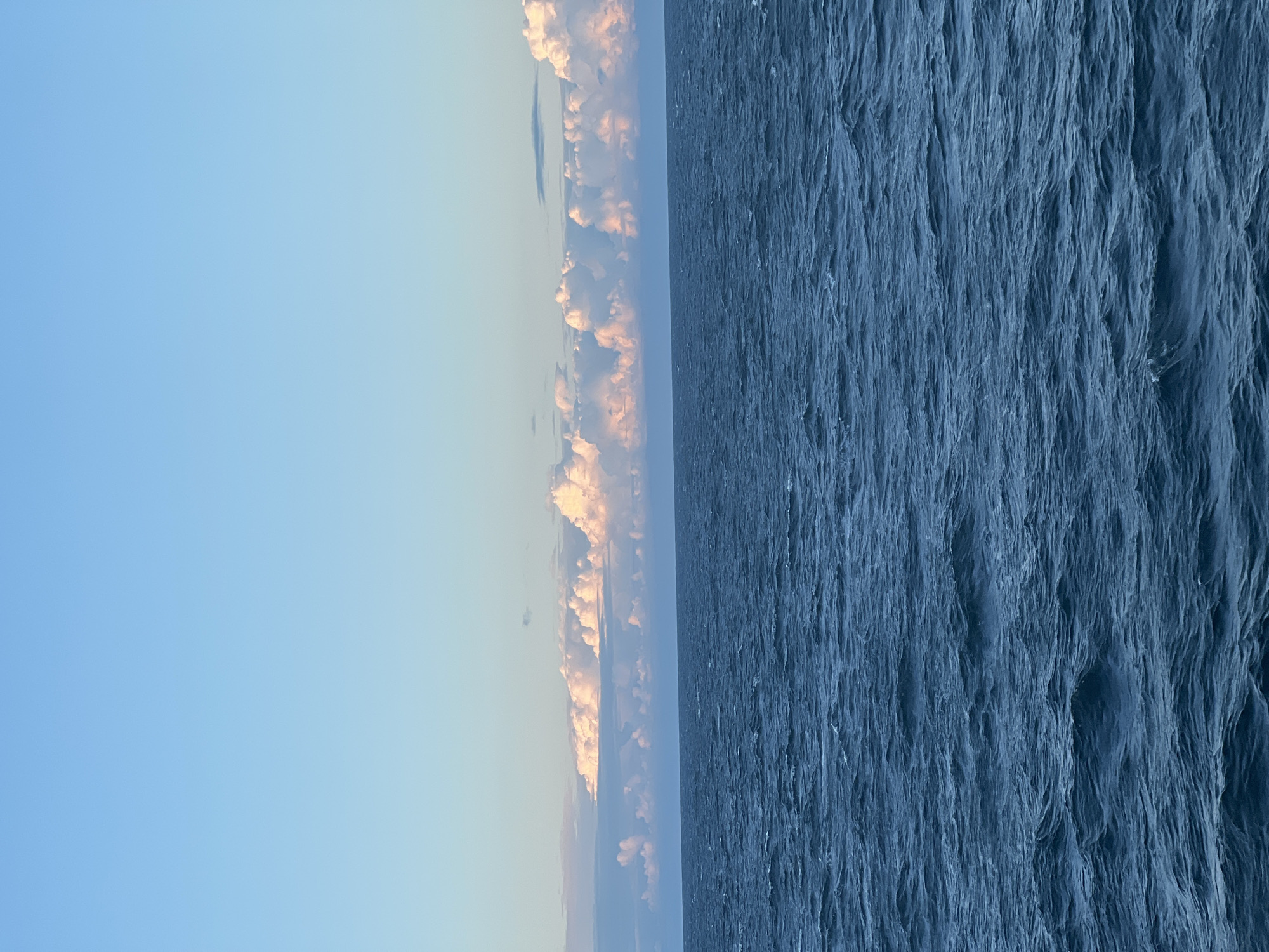 The image depicts a wide expanse of deep blue ocean under a pale blue sky. The water exhibits a choppy texture with small waves. Along the horizon, a band of clouds stretches across the frame. These clouds are illuminated from above with soft pink and white hues, suggesting either sunrise or sunset. The scene is devoid of any people, man-made objects, or distinct geographical landmarks that would indicate a specific location within a city or country.