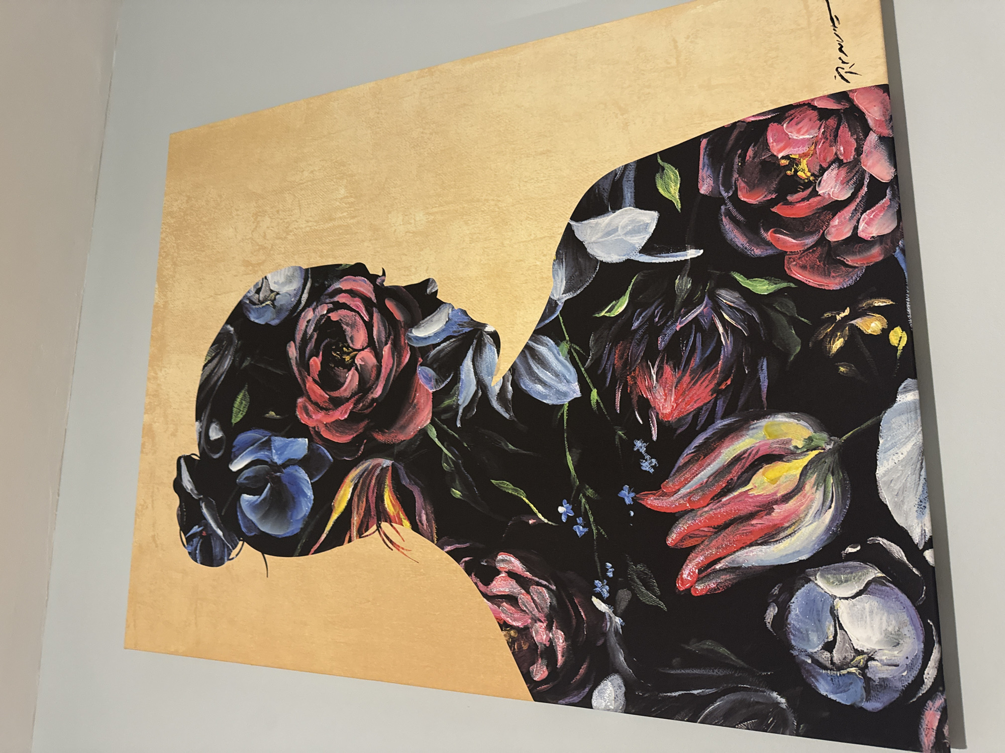 A close-up, eye-level shot depicts an abstract painting. The left portion of the painting is a solid tan color. The right portion is a dark floral arrangement, shaped in the form of a person looking upward. The flowers are various colors, including red, white, blue, and pink. A signature is visible in the upper right corner of the painting. The painting is hung on a light grey wall. The city and location are unknown.