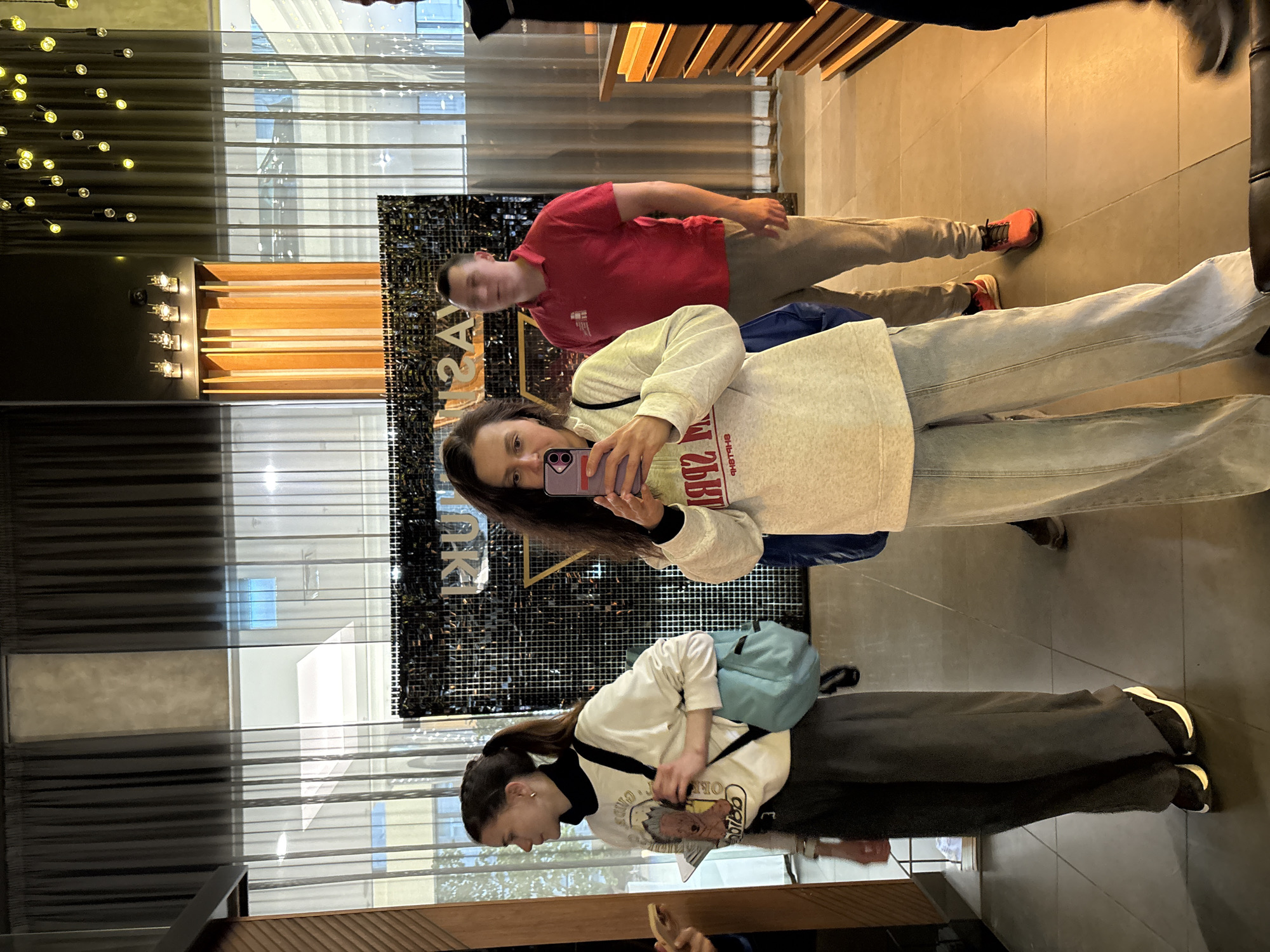 The provided image is oriented sideways, appearing as a landscape view when rotated 90 degrees clockwise. It captures three individuals reflected in a large mirror within a modern indoor setting, likely a public area such as a lobby or hall, in Kazan, Russia.
The setting features a light-colored tiled floor and a dark, textured wall or partition in the background, adorned with shimmering elements. This reflective surface displays reversed text, notably a large "YAS" or similar letters. Overhead, modern lighting fixtures include a decorative chandelier-like element with multiple small, glowing bulbs, and linear recessed lights integrated into a wooden panel. Transparent window-like structures with horizontal blinds are also visible, suggesting some natural light, though the interior is primarily lit by artificial sources.
In the center of the reflection, a young woman with long, dark hair holds a purple smartphone to take a picture, looking directly at the camera with a neutral expression. She wears a light-colored hooded sweatshirt with reversed text on it and light-wash jeans. To her right (as if facing the mirror), a man with short, dark hair is partially visible, wearing a red polo shirt and light-colored trousers with orange sneakers. To the woman's left, another young woman with long, dark hair tied in a ponytail looks downwards at an object in her hands. She is dressed in a white long-sleeved top with a black collar and a graphic print, dark wide-leg trousers, and black and white sneakers, with a light blue crossbody bag slung over her shoulder.
The scene captures a casual moment, possibly a group selfie or photo taken in a large mirror. The time of day is not explicitly clear, but the ambiance suggests an interior space well-lit by modern fixtures.