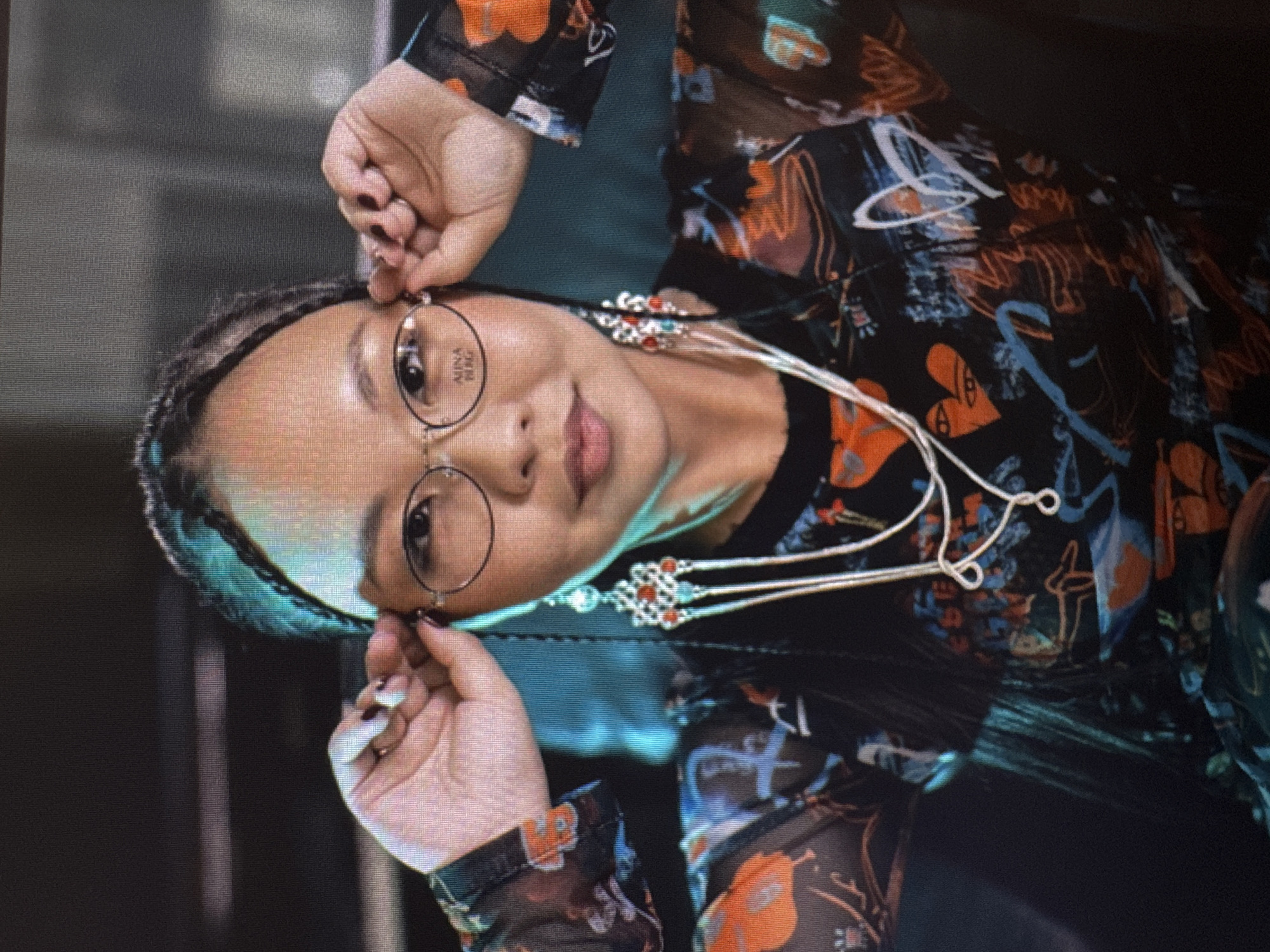 The image is a close-up shot of a person, likely a woman, wearing glasses and elaborate earrings. She is looking towards the camera and adjusting her glasses with her hands. Her hair is braided with some blue-green streaks, and she is wearing a dark, sheer top with an orange and blue pattern. The background is dark and out of focus, suggesting an indoor setting. There is no visible information in the image that would indicate the location in Irkutsk, Russia.