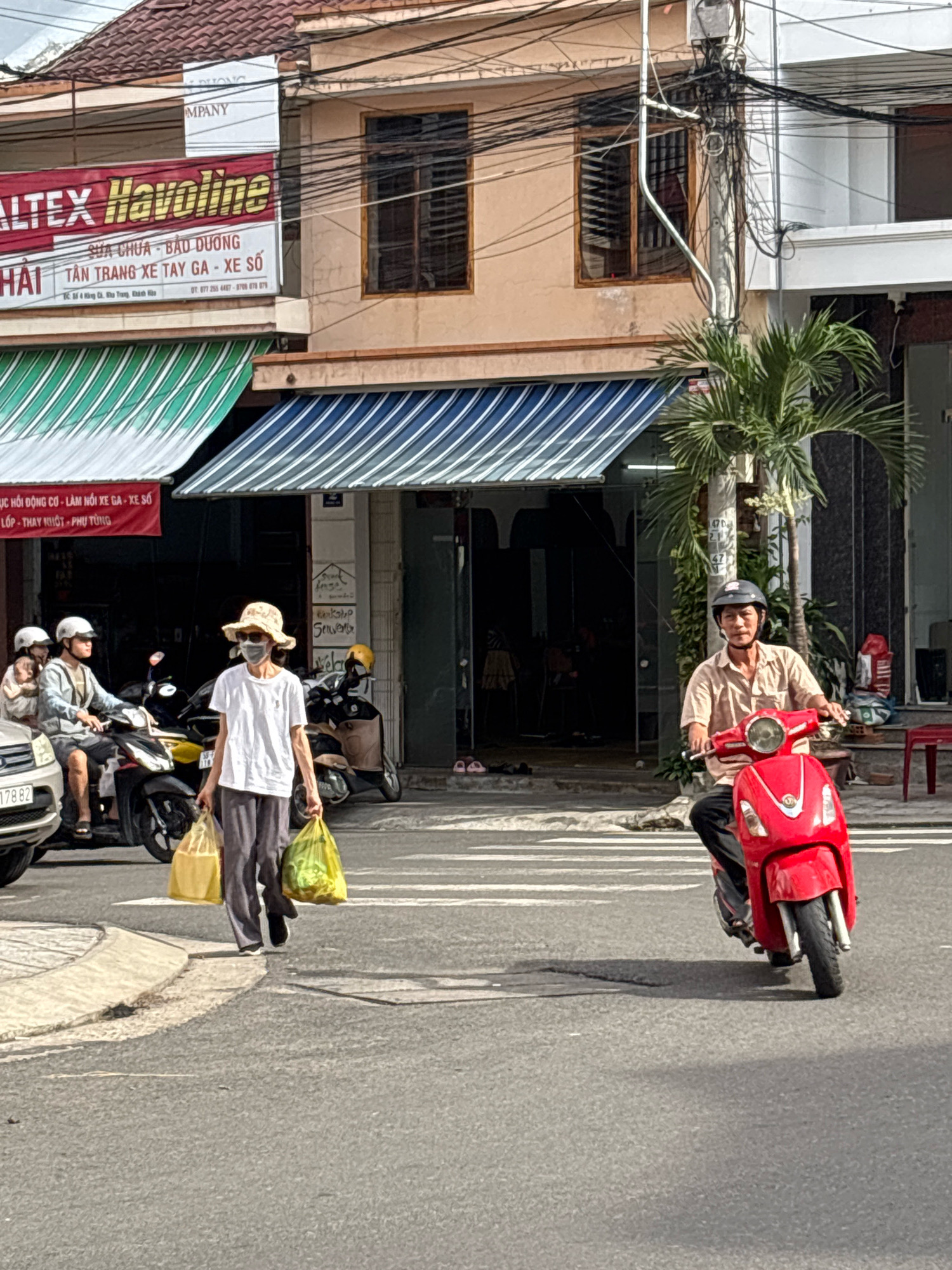 The image captures a street scene in Nha Trang, Vietnam, likely on a sunny day, given the bright lighting and shadows. In the foreground, a man wearing a light-colored shirt, dark pants, and a helmet rides a red scooter. He is looking forward, his hands on the handlebars. To the left, a woman walks across a pedestrian crossing, carrying two shopping bags, one yellow and one green. She is wearing a white t-shirt, dark pants, a straw hat, and a face mask. She is looking down. Behind her, a person on a motorcycle, also wearing a helmet, is carrying a child. The motorcycle has a white license plate that is partially visible. The background features buildings with awnings, some of which have striped patterns in green and white, and blue and white. There are power lines and poles visible, typical of urban Vietnamese streetscapes. The overall impression is of everyday life unfolding on a busy street.