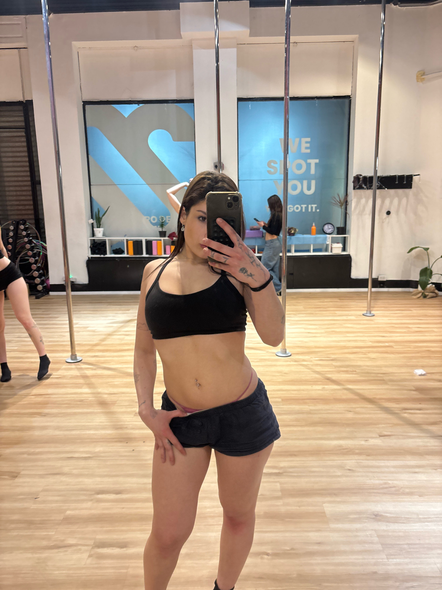 The image depicts a person taking a mirror selfie in what appears to be a dance or fitness studio. The setting includes several tall, metallic poles, a wooden floor, and large windows with blue and white graphics. In the background, another person can be seen. The studio is equipped with shelves holding various items, possibly for classes. The overall ambiance suggests a modern, active space. The location is not specified as Buenos Aires, Argentina.