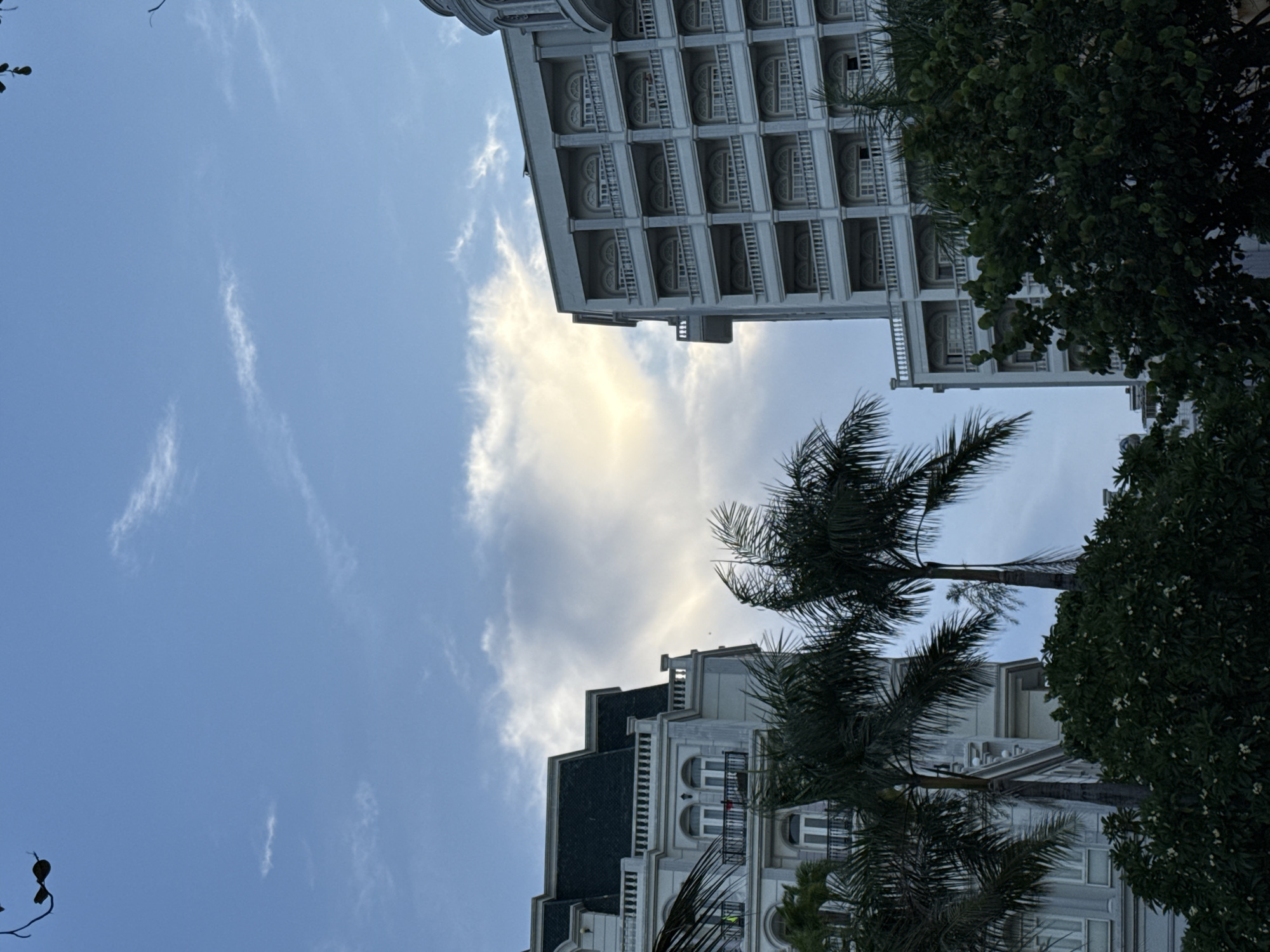The image, taken in Nha Trang Ward, Vietnam, showcases a low-angle view of two ornate white buildings against a backdrop of a bright blue sky with wispy white clouds. Palm trees with their characteristic fronds are visible in the foreground and between the buildings, suggesting a tropical or subtropical setting. The architectural style of the buildings appears to be classic, with detailed facades, multiple balconies, and arched windows. The lighting indicates it is daytime, likely morning or late afternoon, given the softness of the light. There are no people or specific activities depicted; the focus is on the architecture and the natural elements. The scene evokes a sense of tranquility and vacation, typical of a resort area.