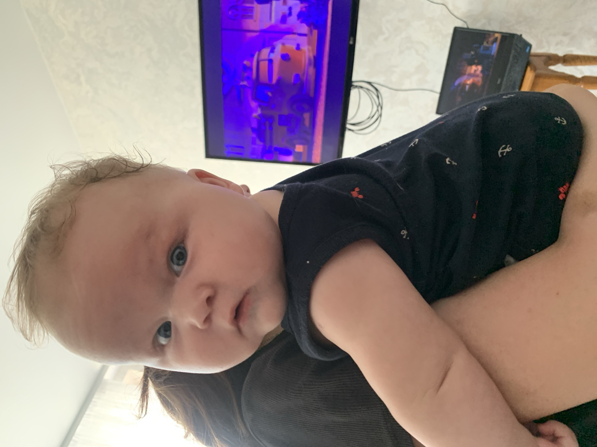 A baby is held in someone's arms, looking directly at the camera. The baby has blonde curly hair and blue eyes. They are wearing a dark blue onesie with white anchors on it. In the background, a television screen displays a bright purple image, and a computer tower with colorful lights is visible on a table. The scene appears to be indoors, likely a living room, with a textured wall and ceiling. The overall lighting suggests it could be daytime. The baby's expression is neutral and curious. The image is oriented sideways.