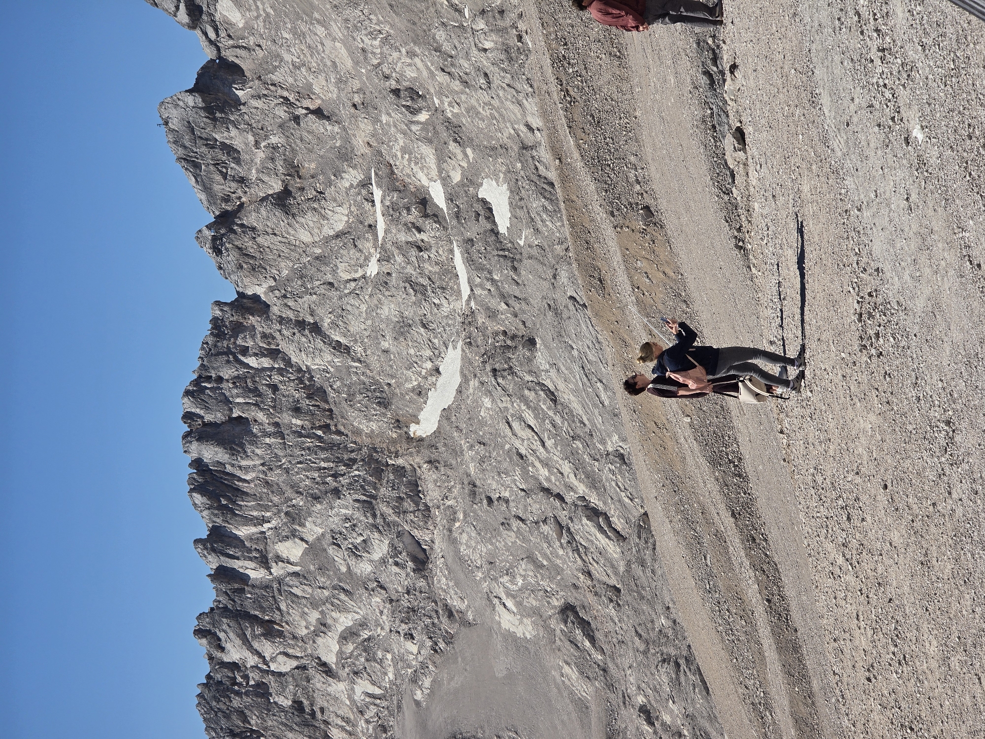 The image captures a panoramic view of a rugged, rocky mountain landscape under a clear blue sky. The foreground features a gravelly path with a few scattered rocks and sparse vegetation. Two individuals are standing on the path in the middle ground, with one person appearing to take a photo with their phone. A third person is visible further to the right, walking away from the camera. The background is dominated by the imposing, grey, and textured rock face of a mountain range, with patches of snow visible on some of the higher slopes. The overall scene depicts a remote, high-altitude environment. This location is likely the Zugspitze, Germany's highest peak, which is accessible from Garmisch-Partenkirchen via a cable car or cogwheel train.