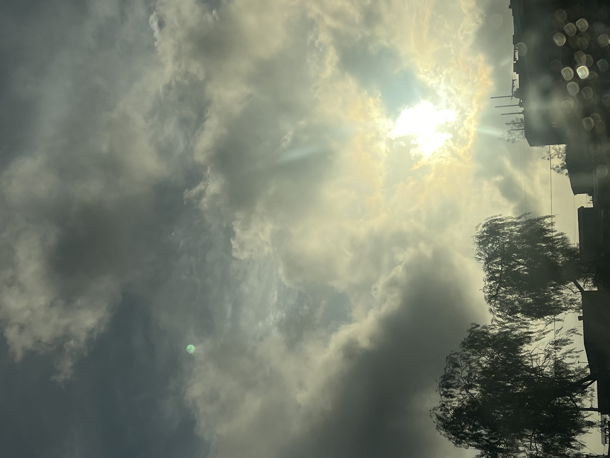 The image shows a sky with dense, layered clouds obscuring the sun in Arica, Chile. The sun peeks through a gap in the clouds, creating rays of light. Two trees with visible wind-blown leaves are on the lower right, adjacent to a building's edge, with antennas on the roof. Bokeh effect creates blurred circles in the right foreground. A green spot appears within the dark clouds on the left.
