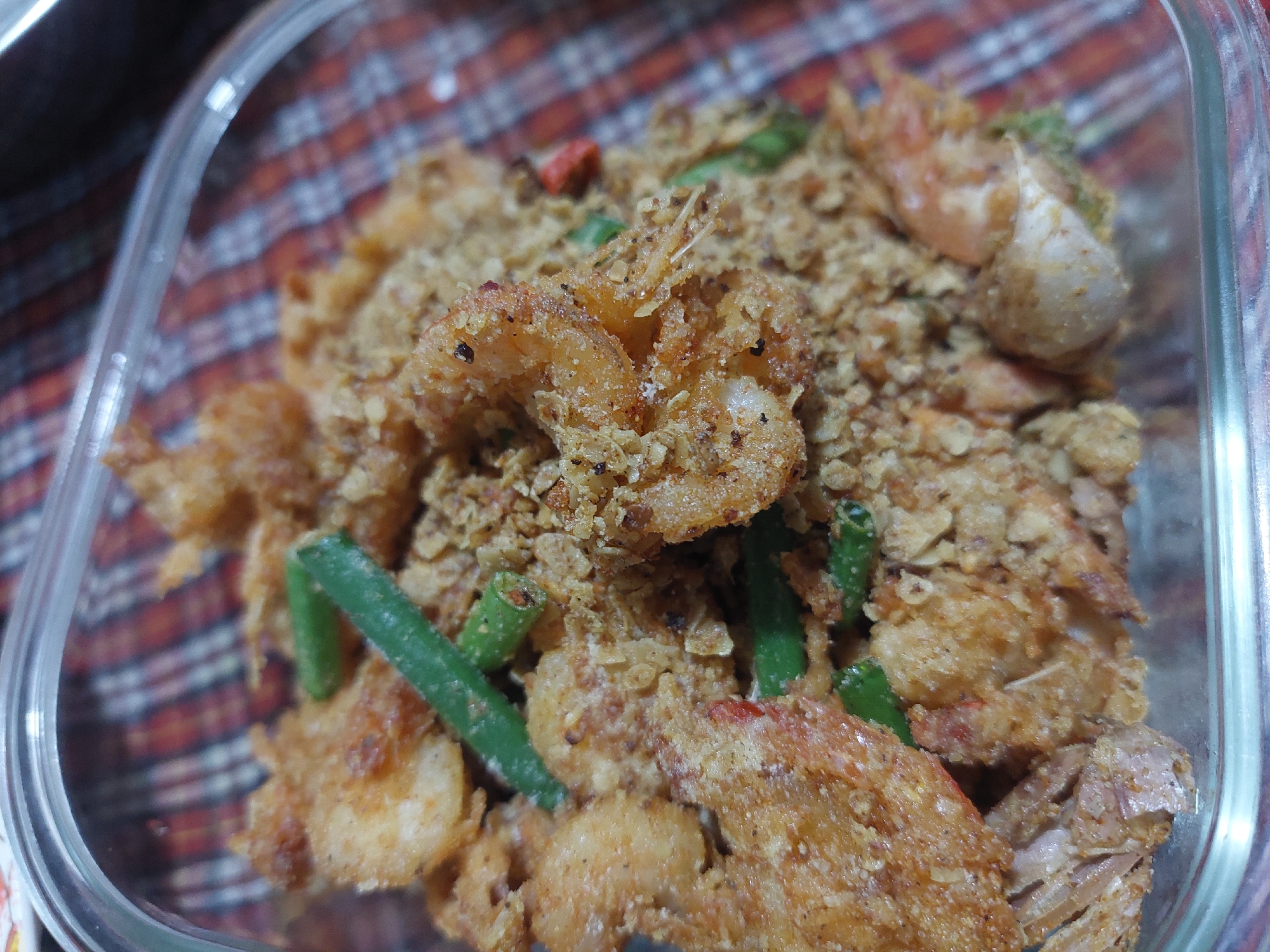 In a clear glass container, a dish of fried shrimp and green beans is presented. The shrimp are coated in a crispy, golden-brown batter and mixed with small, finely ground pieces of what appears to be nuts or breadcrumbs, giving the dish a textured appearance. Several vibrant green beans are interspersed throughout the shrimp. The container is placed on a surface covered with a red and white plaid fabric. The lighting suggests an indoor setting, possibly a home kitchen or a restaurant. No people are visible in the image, and no specific location in Jelutung, Indonesia, can be determined.