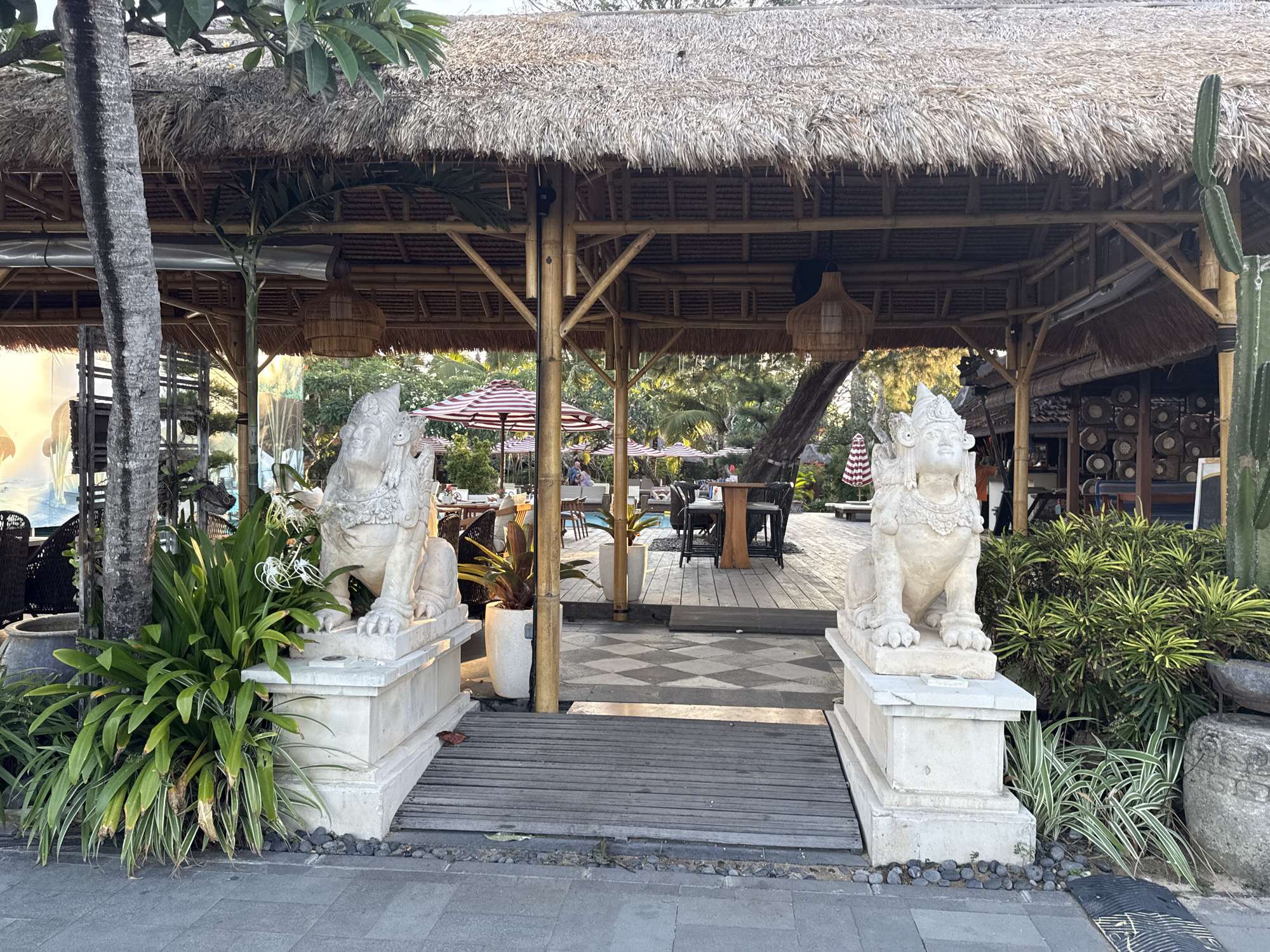 This image depicts an outdoor seating area of a restaurant or resort, likely located in Denpasar, Indonesia. The scene is characterized by a thatched-roof structure supported by bamboo poles, creating an open-air dining space. Two stone lion statues stand guard at the entrance to this area, flanking a wooden walkway. Lush green foliage, including palm trees and other tropical plants, surrounds the statues and the seating area. In the background, several tables and chairs are visible, some with red and white striped umbrellas, suggesting a sunny and inviting atmosphere. A pool can also be seen in the distance. The overall impression is one of a tropical, relaxed ambiance, typical of beachside or resort locations in Bali.