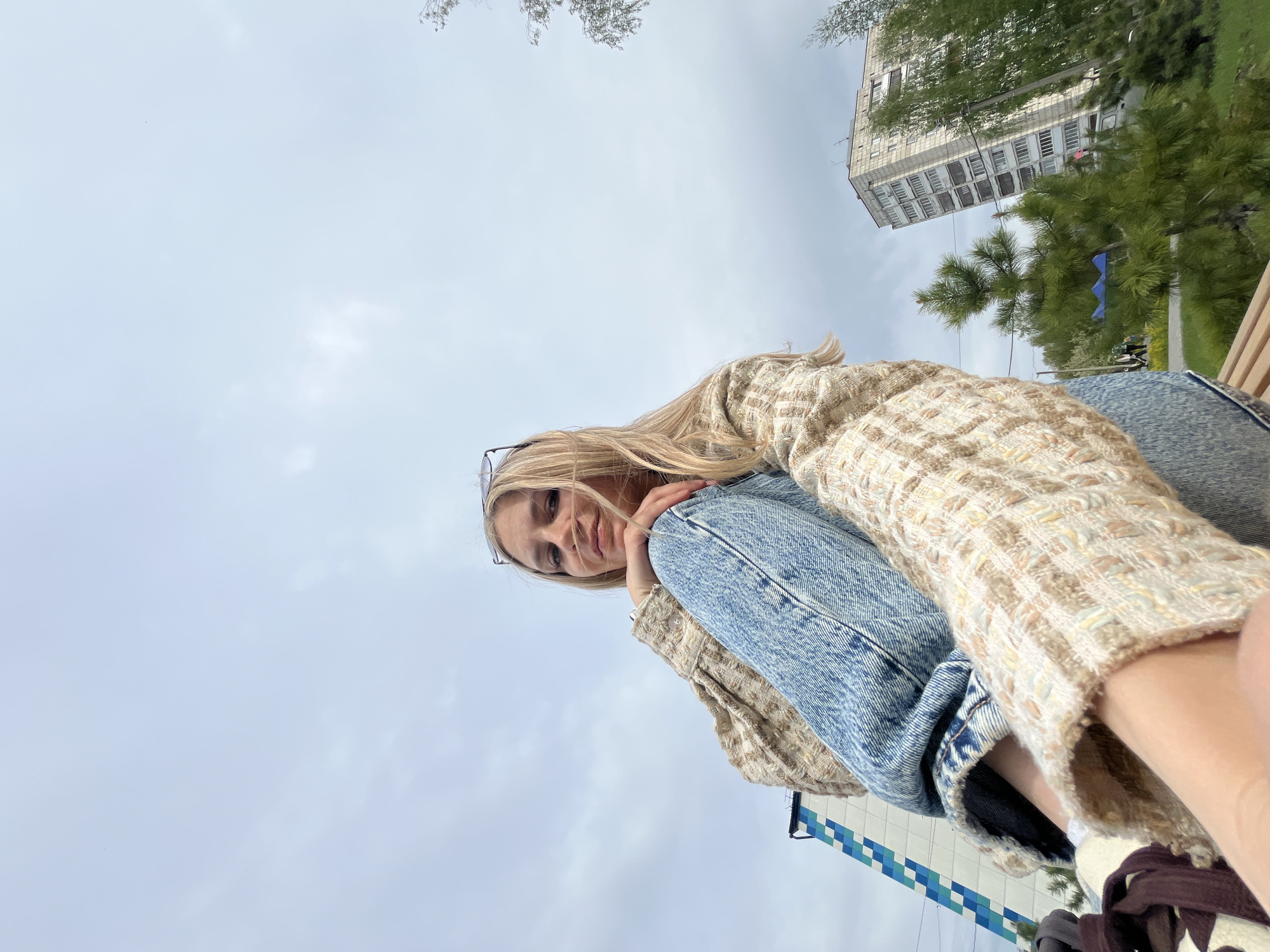 A woman is sitting outdoors, possibly on a bench, with a view of the sky, buildings, and greenery. She is wearing jeans and a textured jacket. The background includes a tall building and trees, suggesting a city environment. The image was taken in Chaikovsky, Russia.